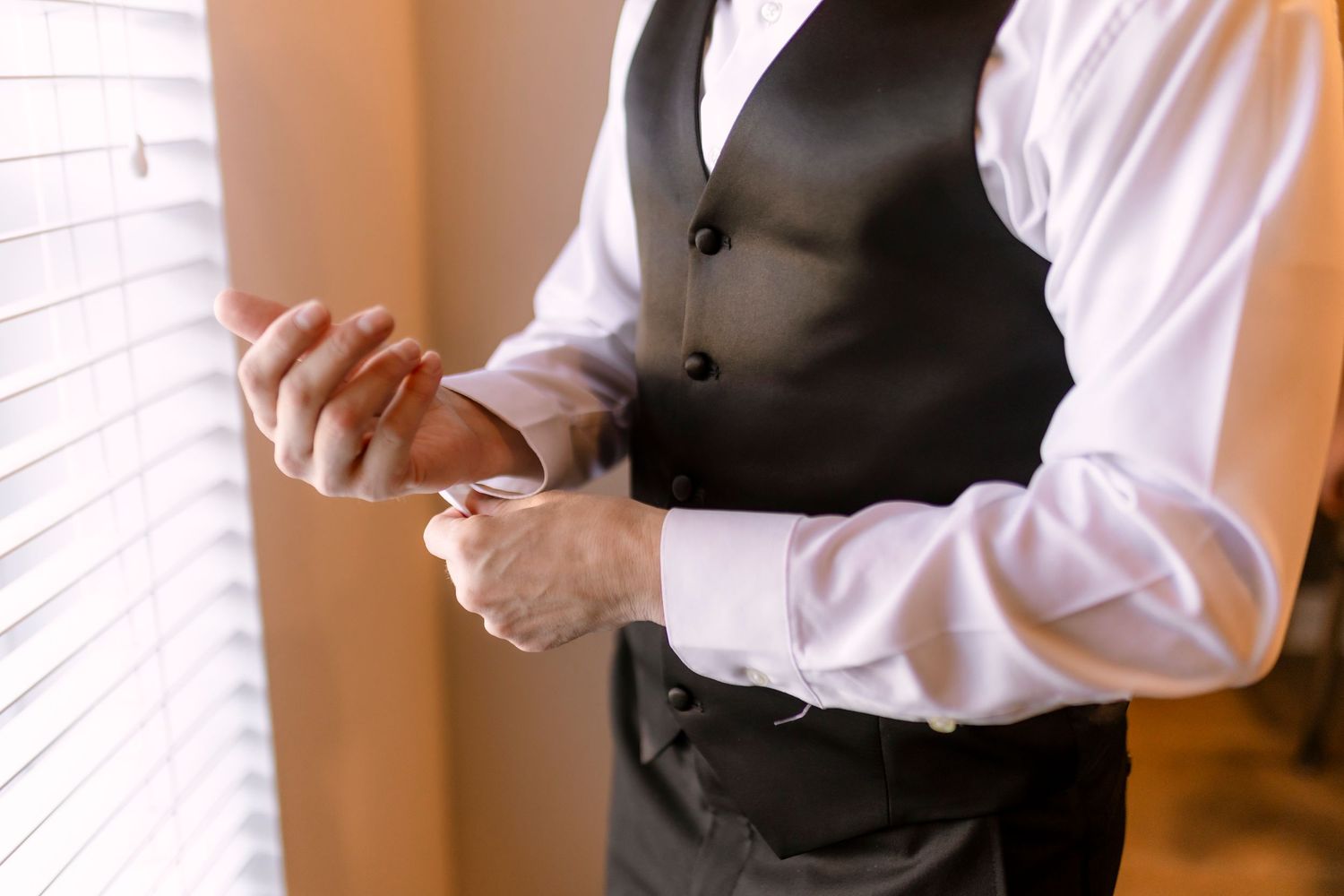 Professional wearing a black vest and white dress shirt adjusts their cuffs while standing near a window.