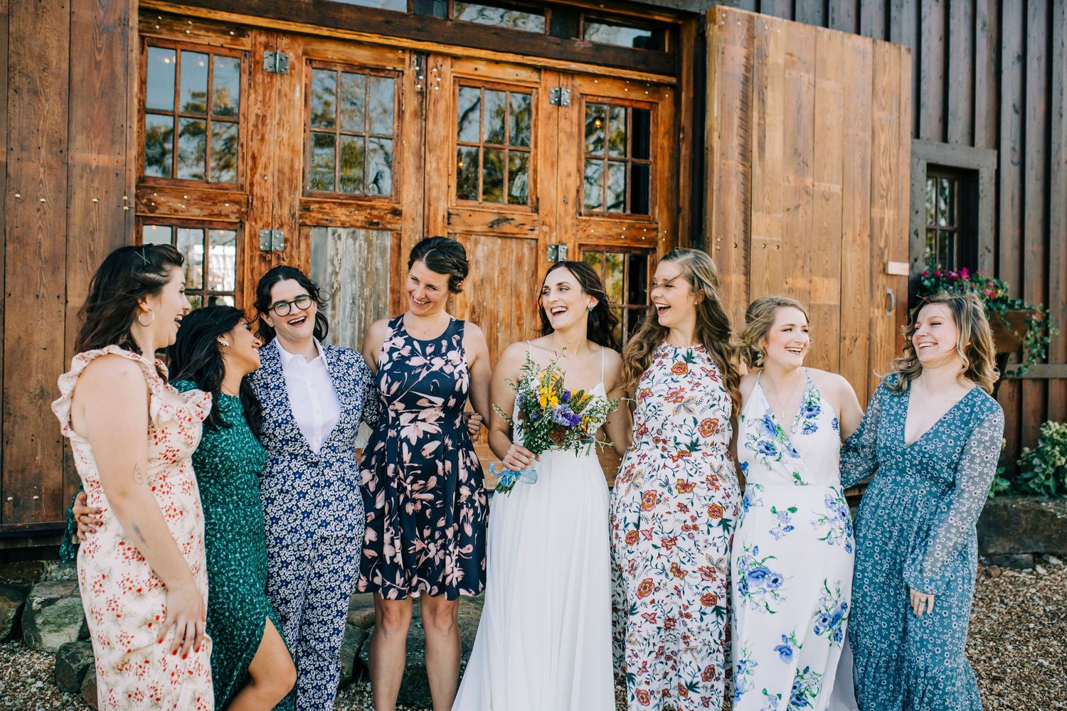 Bridesmaids in floral dresses gather around bride outside wooden barn entrance.