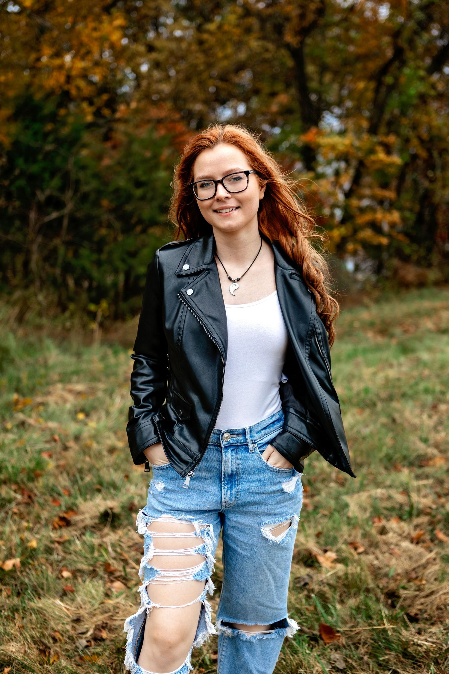 Person in black leather jacket and distressed jeans poses outdoors in autumn with fall foliage in background.
