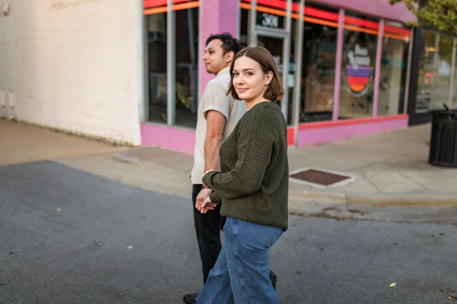 Two people stand casually on an urban street corner with a pink storefront in the background.
