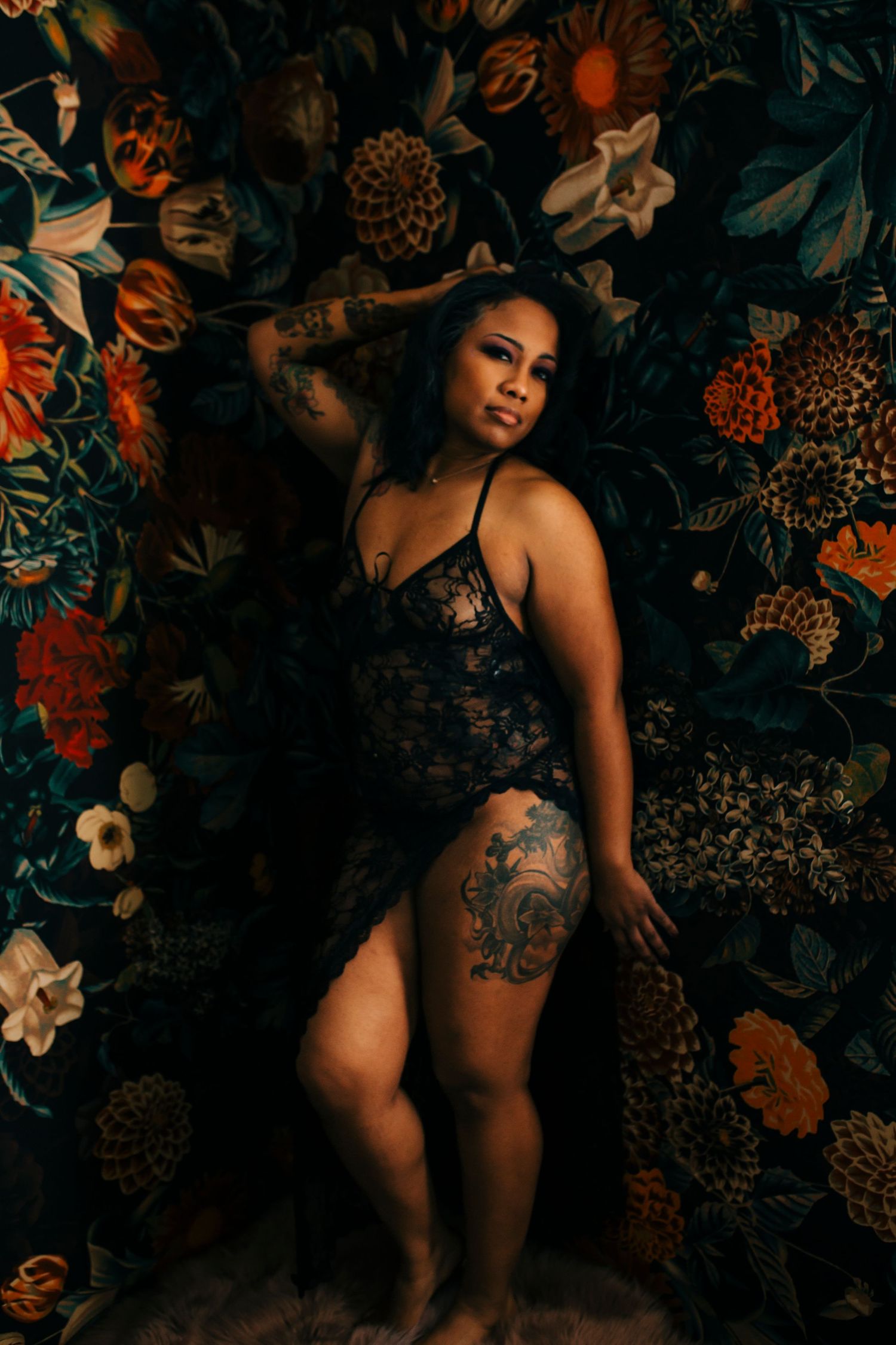 Moody portrait against floral backdrop features dark lace lingerie and visible tattoo art in dramatic lighting.