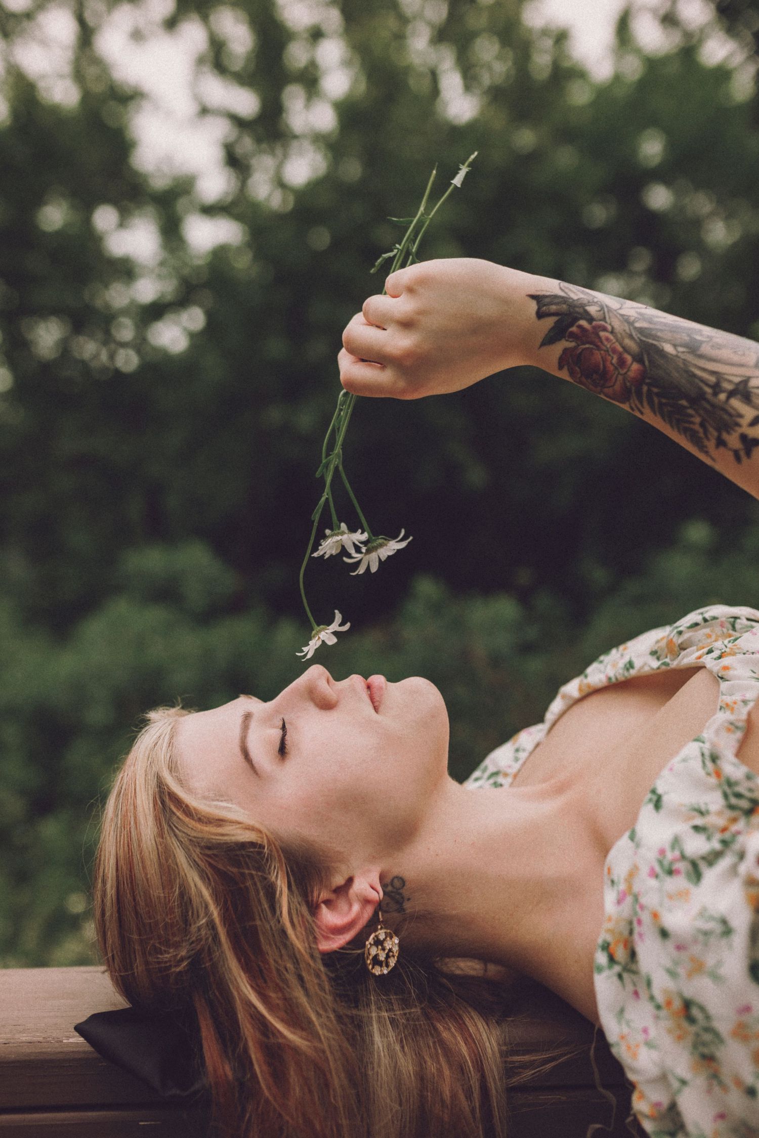 A tattooed arm holding wildflowers over someone lying on a wooden bench in a floral dress against a blurred green background.