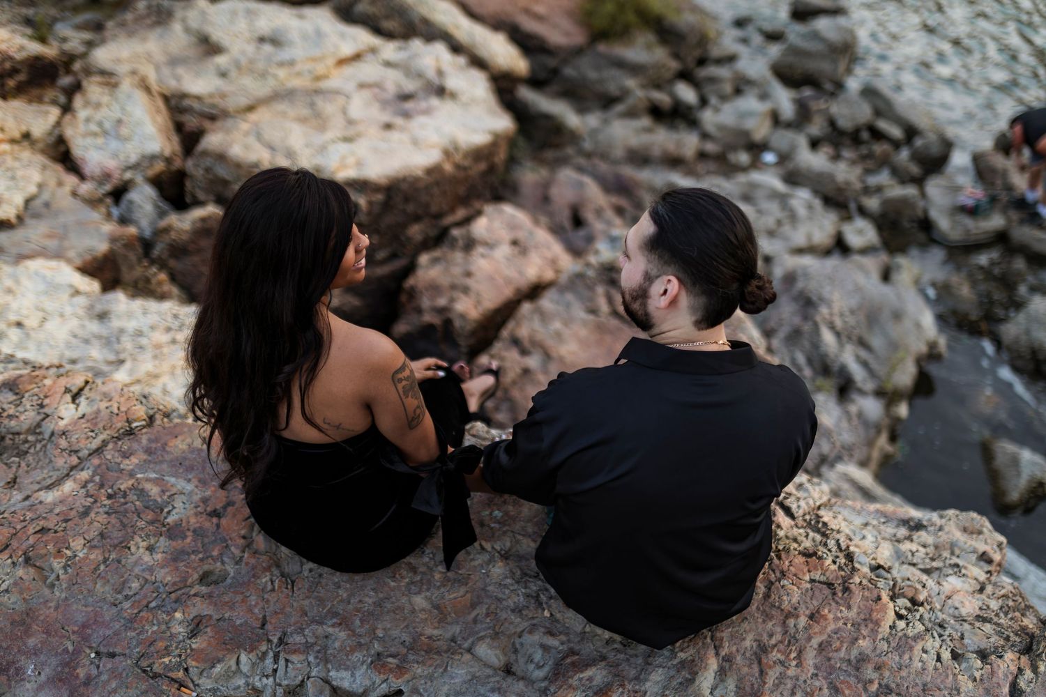 A couple sits together on rocky terrain near a waterfall, enjoying a peaceful moment in nature.