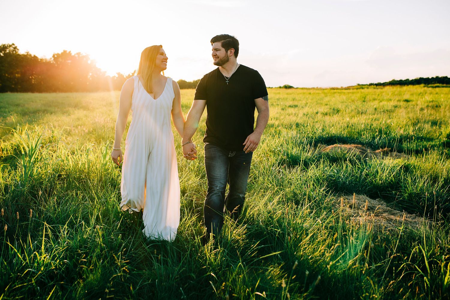 Two people stand together in a sunlit field with tall green grass during golden hour.