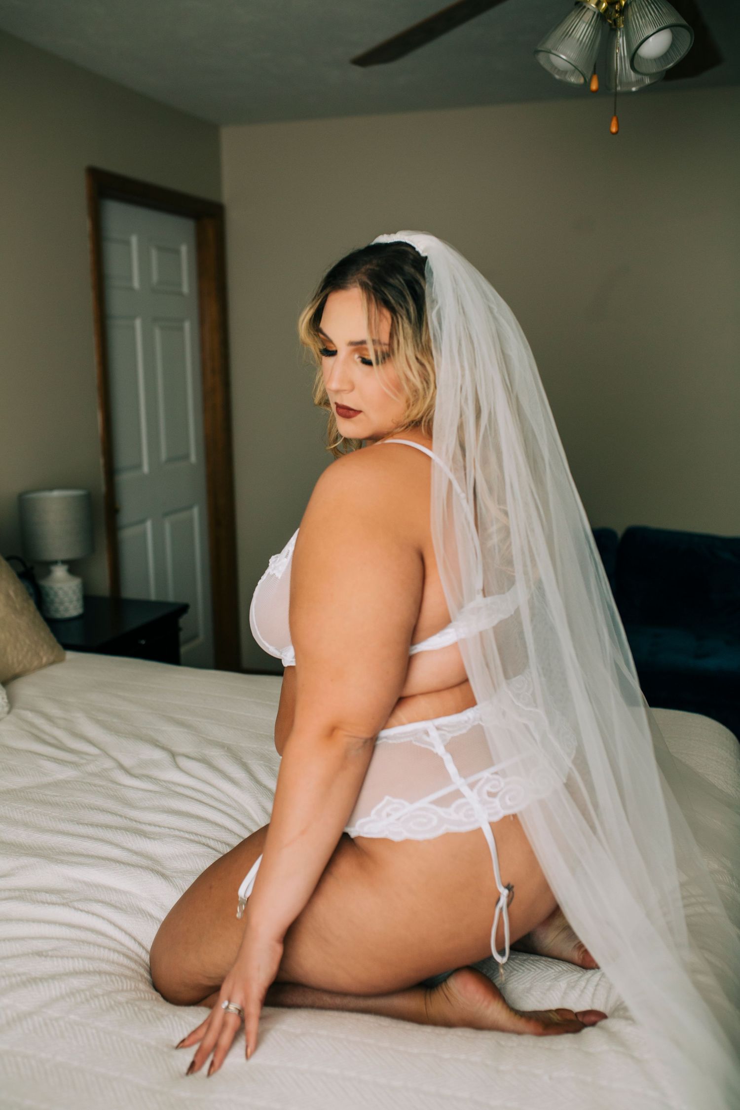 A person wearing white lingerie and a long bridal veil poses on a bed in a bedroom setting.