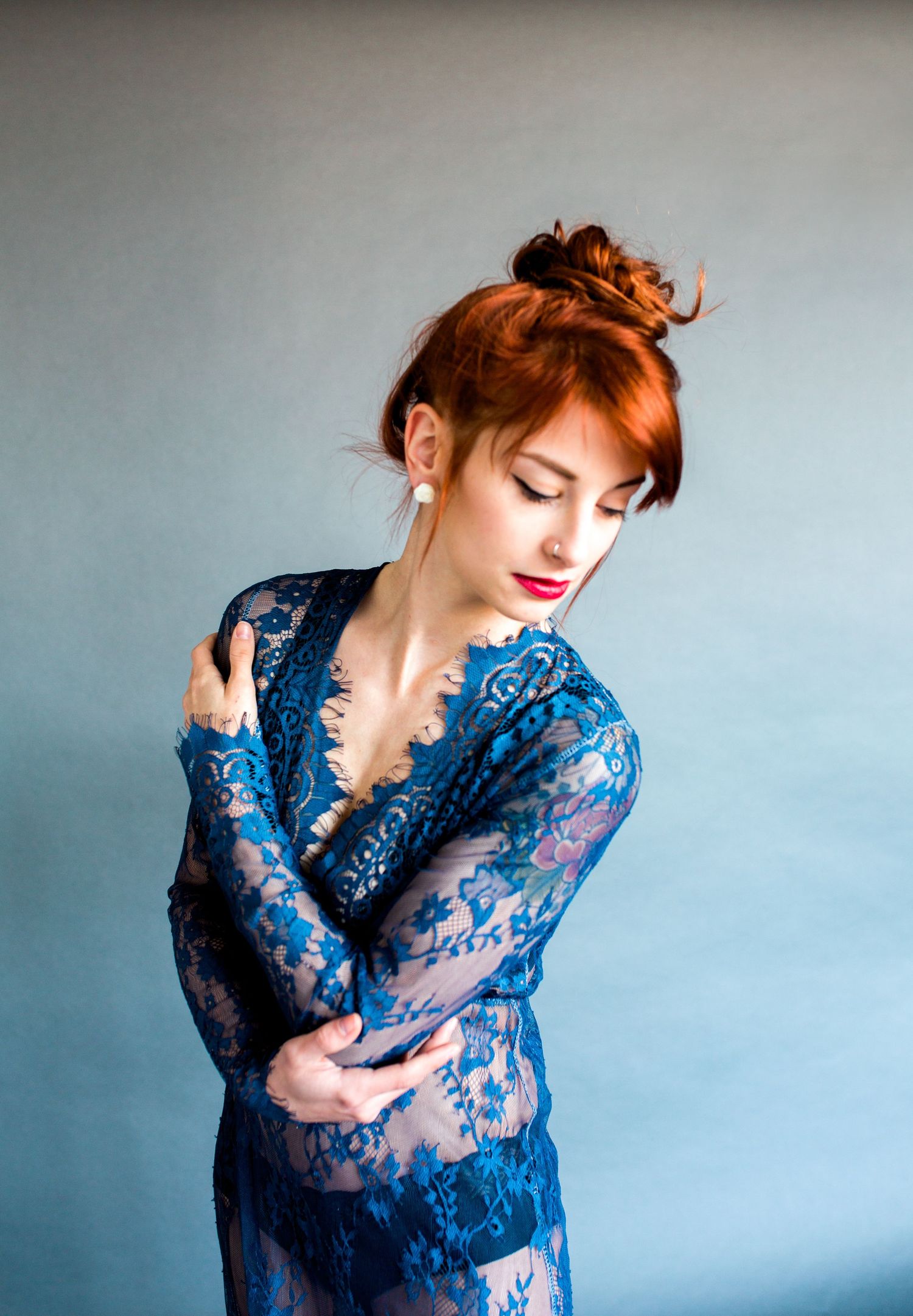 Someone posing in a sheer blue lace robe with their arms wrapped around themselves against a gray backdrop.