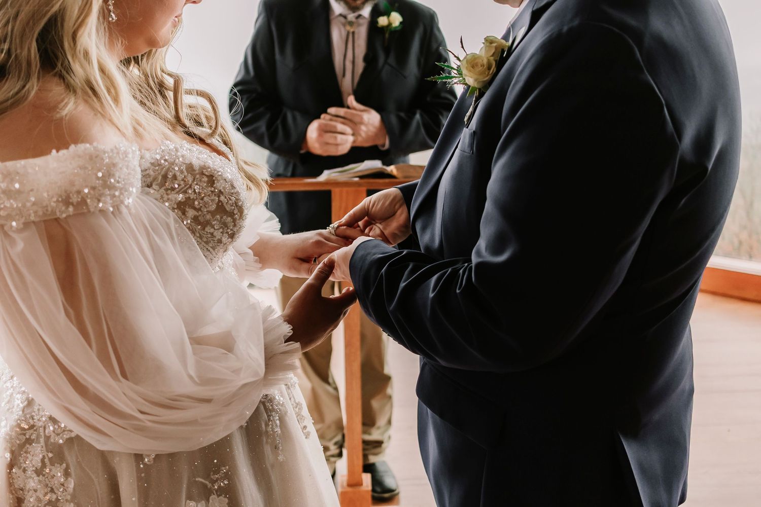 Two people exchange rings during a wedding ceremony while an officiant holds paperwork in the background.