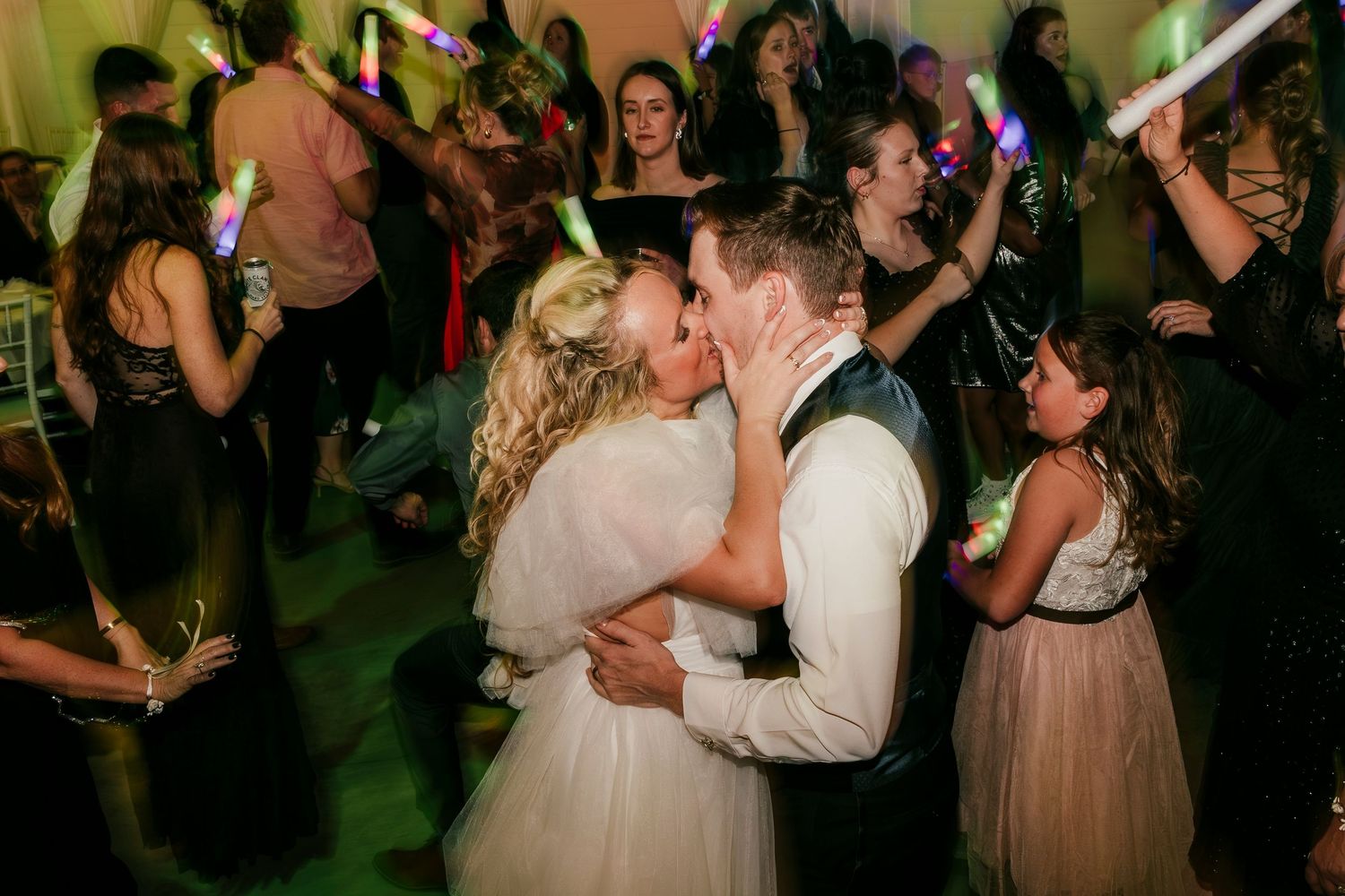 Couple shares romantic kiss on dance floor at wedding reception with crowd dancing in background under green lighting.