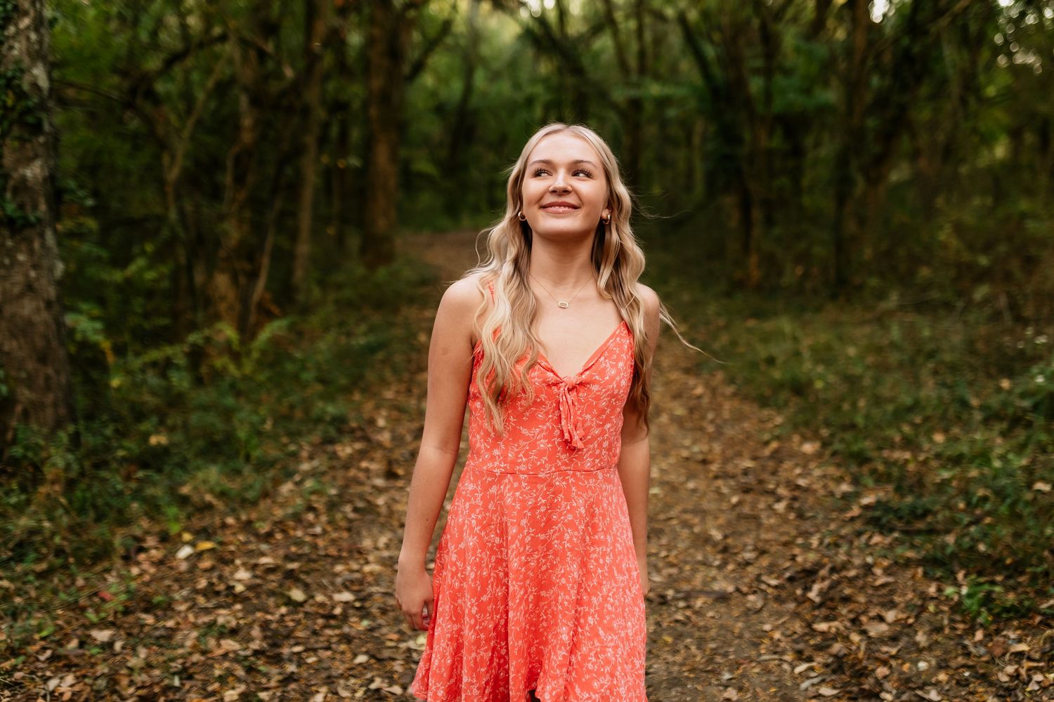 Woman in a coral sundress smiles while standing on a wooded path surrounded by green trees.