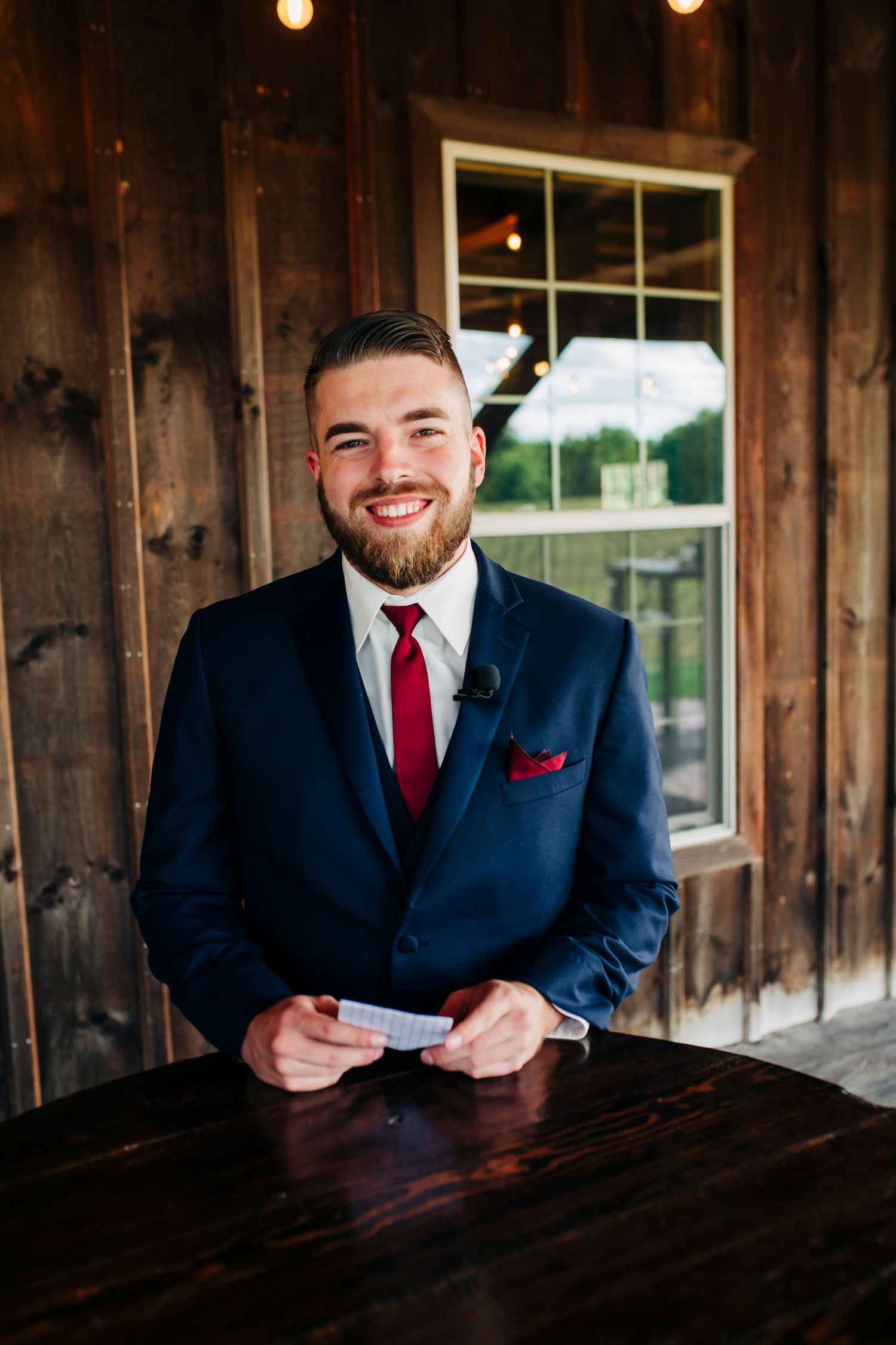 A row of groomsmen in navy suits with red ties pose in a rustic wooden venue.