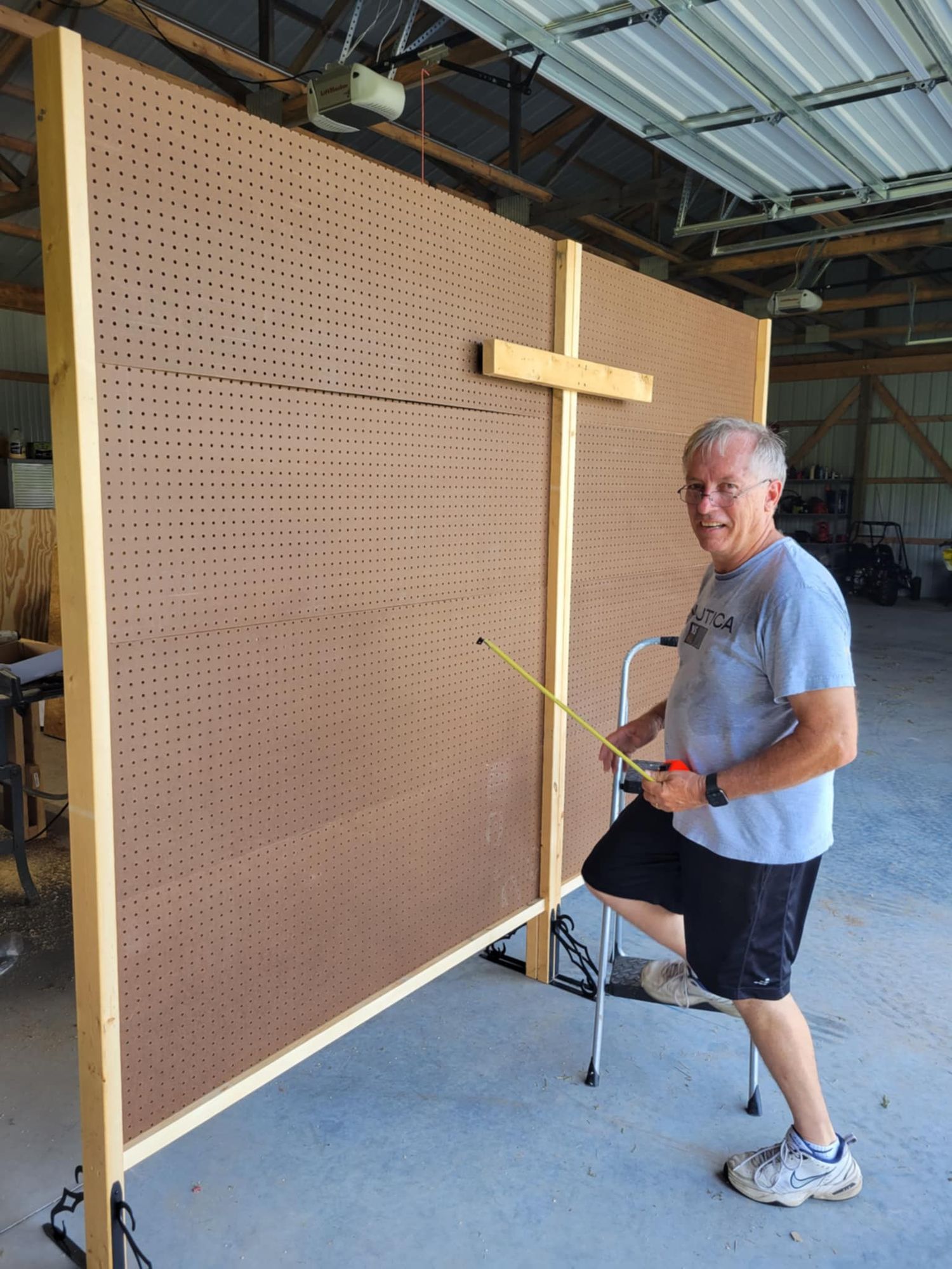 A person stands beside a large pegboard wall with a wooden cross mounted in what appears to be a workshop setting.