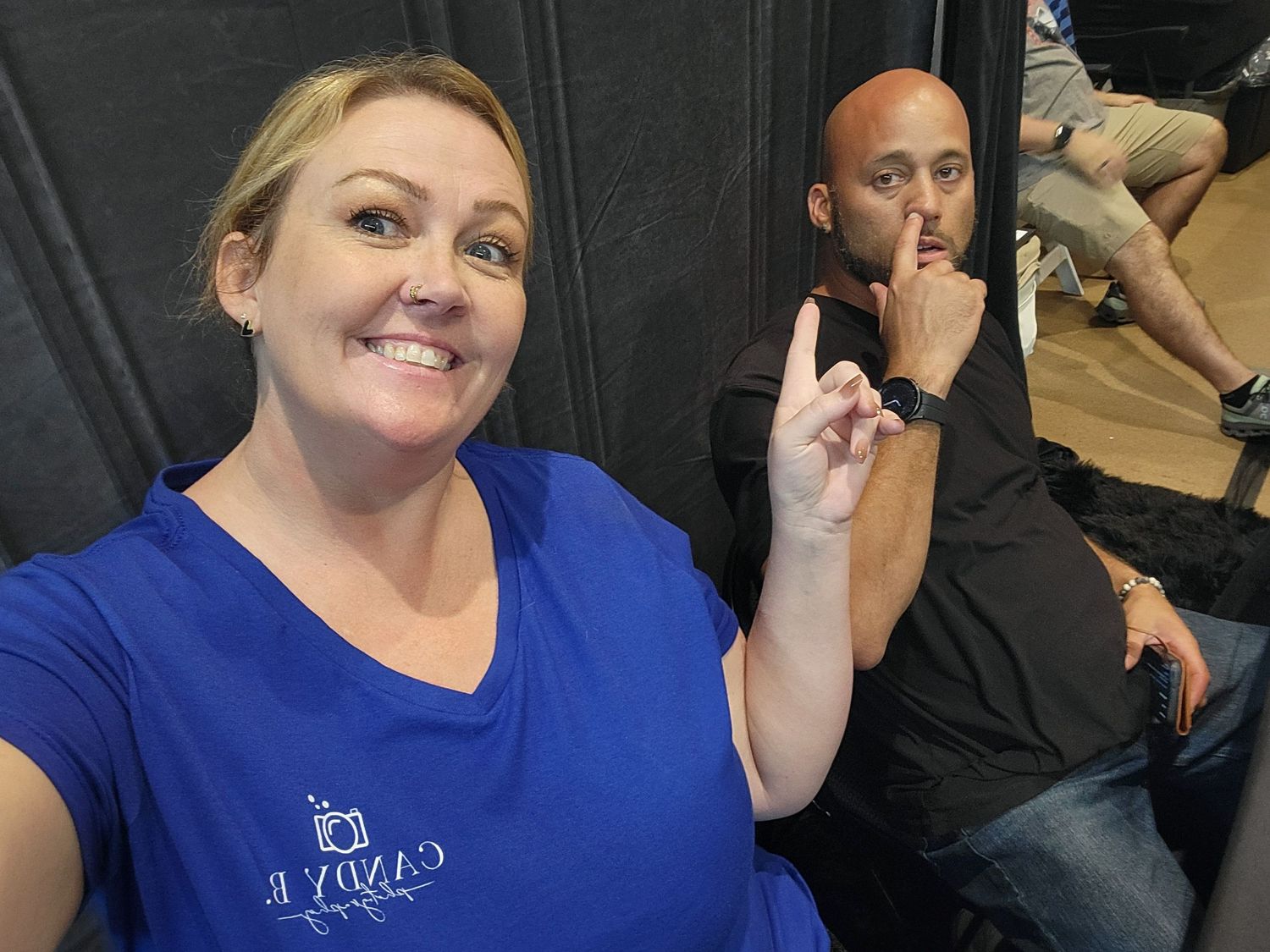 Two people posing in front of a black backdrop, with one wearing a bright blue shirt and gesturing with a finger up.