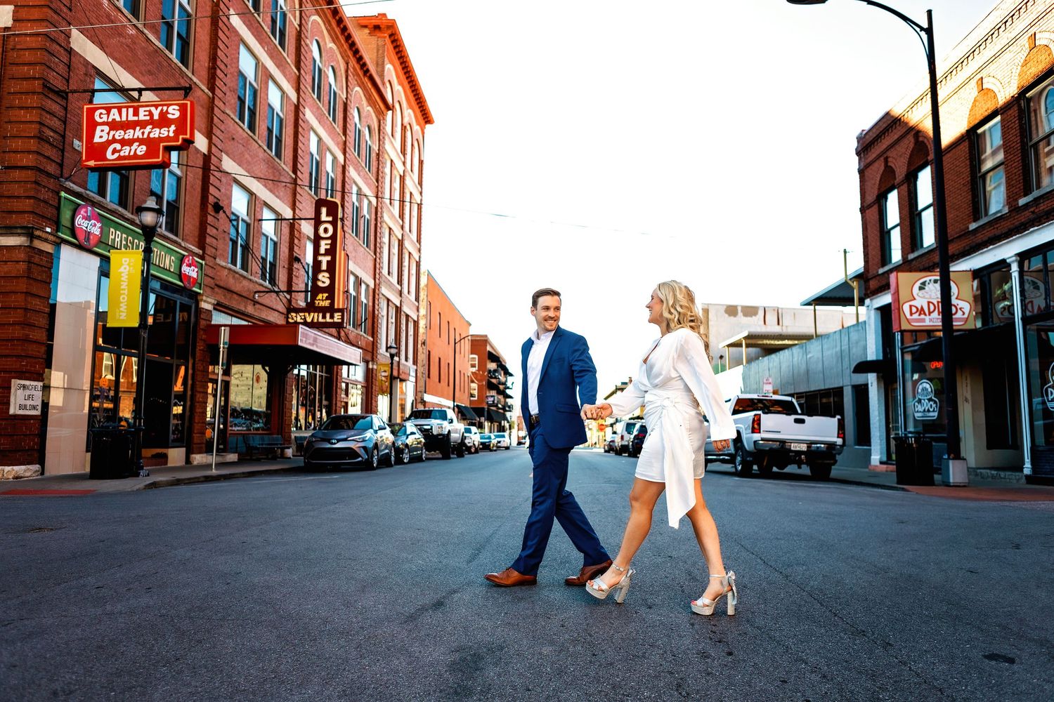 People stroll down a historic brick-paved street with vintage storefronts and commercial buildings in a downtown area.
