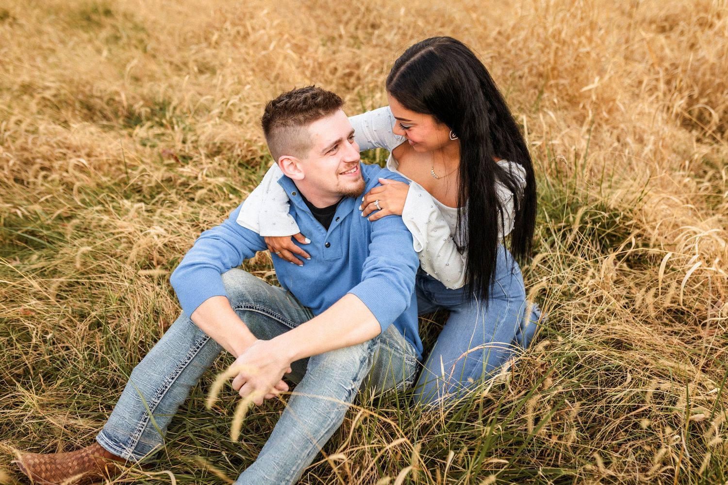 Young couple in matching denim sits intimately together in a golden grass field on a sunny day.