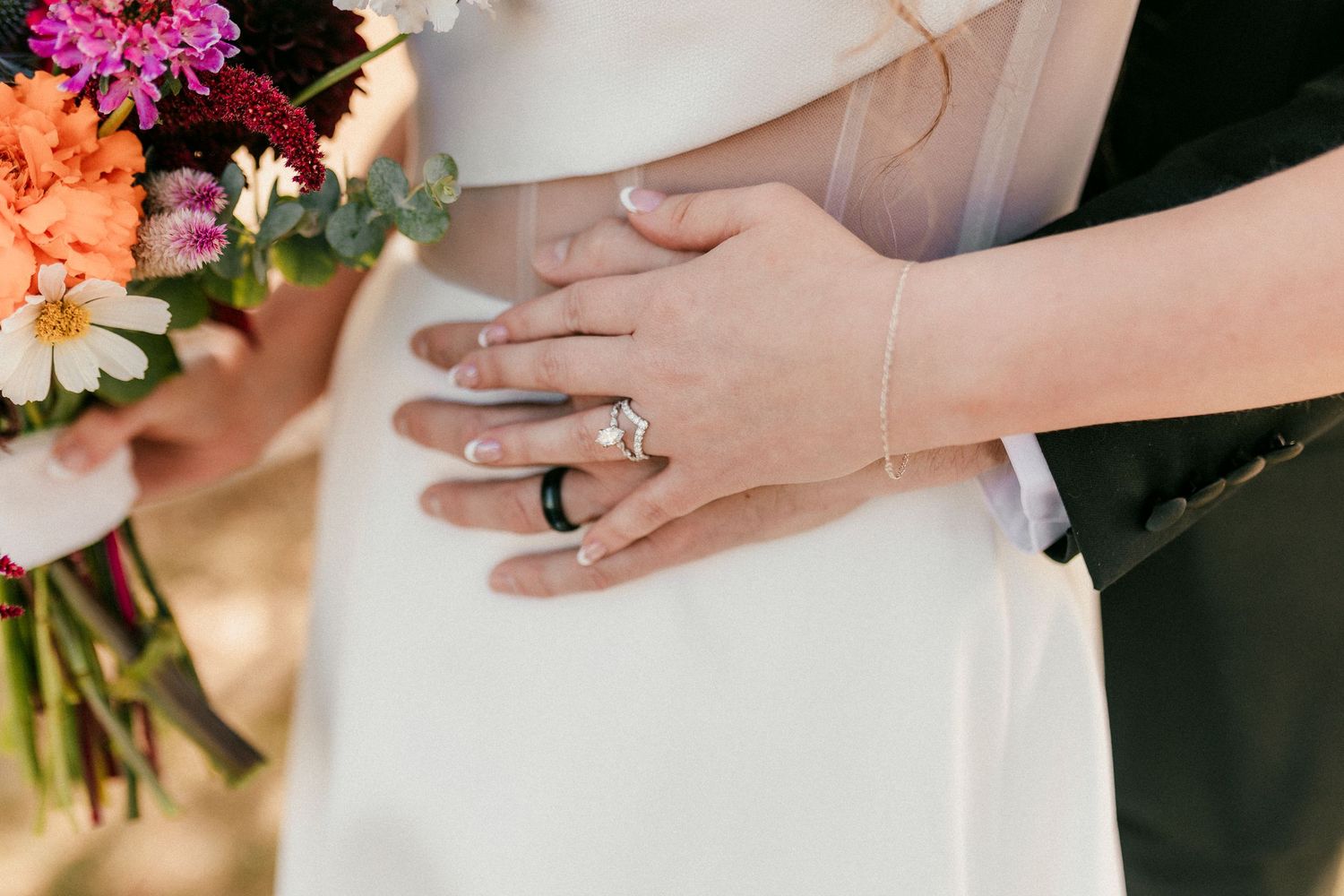 Close-up of hands showing engagement ring with colorful wedding bouquet of pink and peach flowers against white dress.