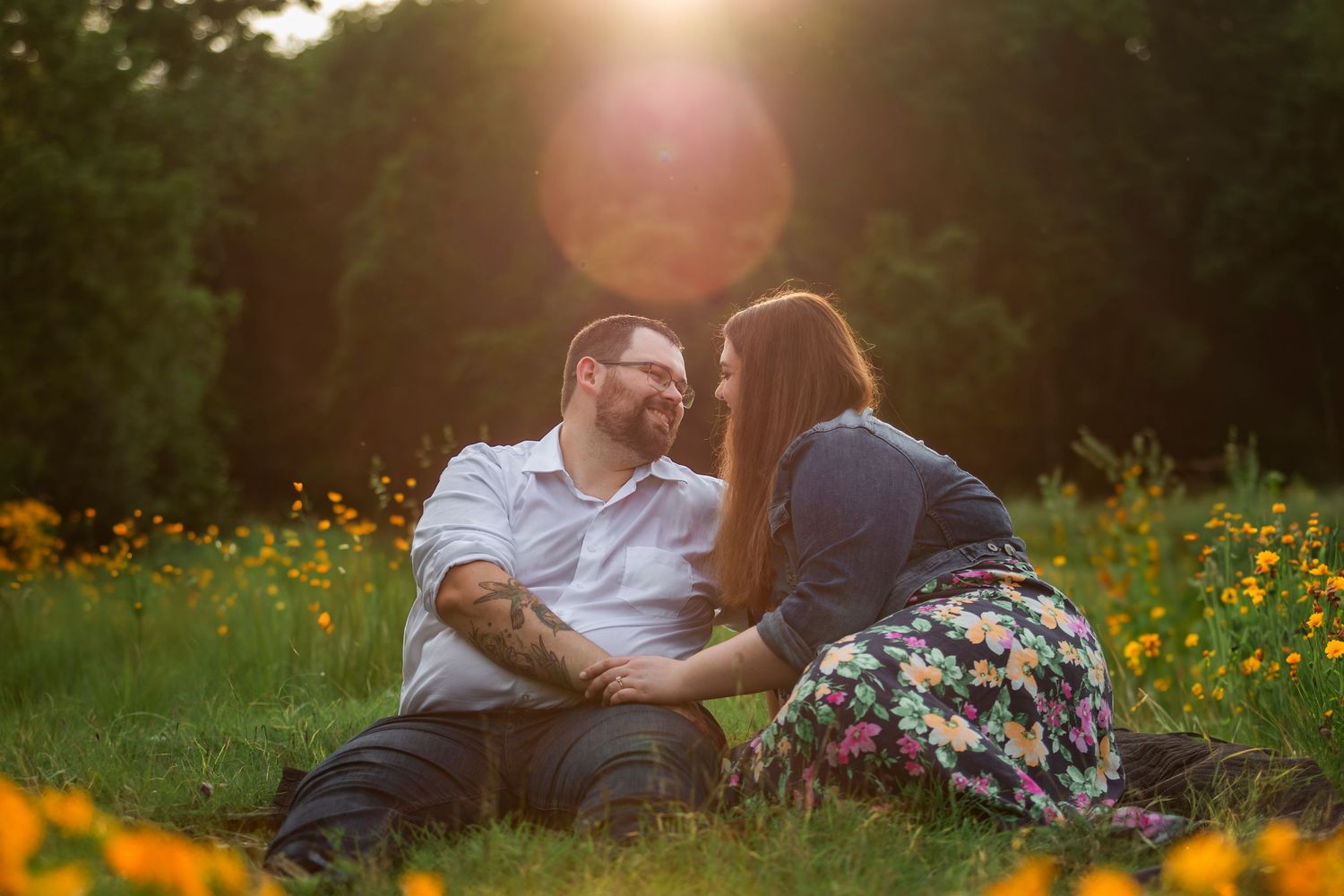 A couple sits together in a sunlit meadow filled with vibrant yellow wildflowers at golden hour.