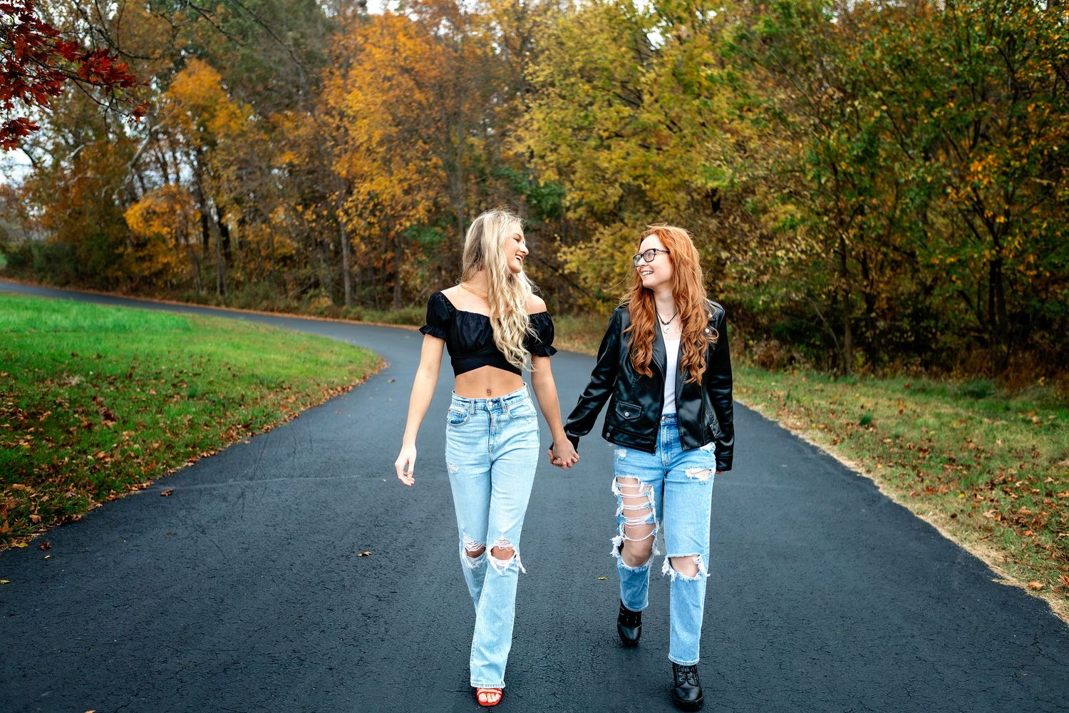 Two sisters in matching light denim jeans walking together on a tree-lined autumn road in Sprinfield, MO.