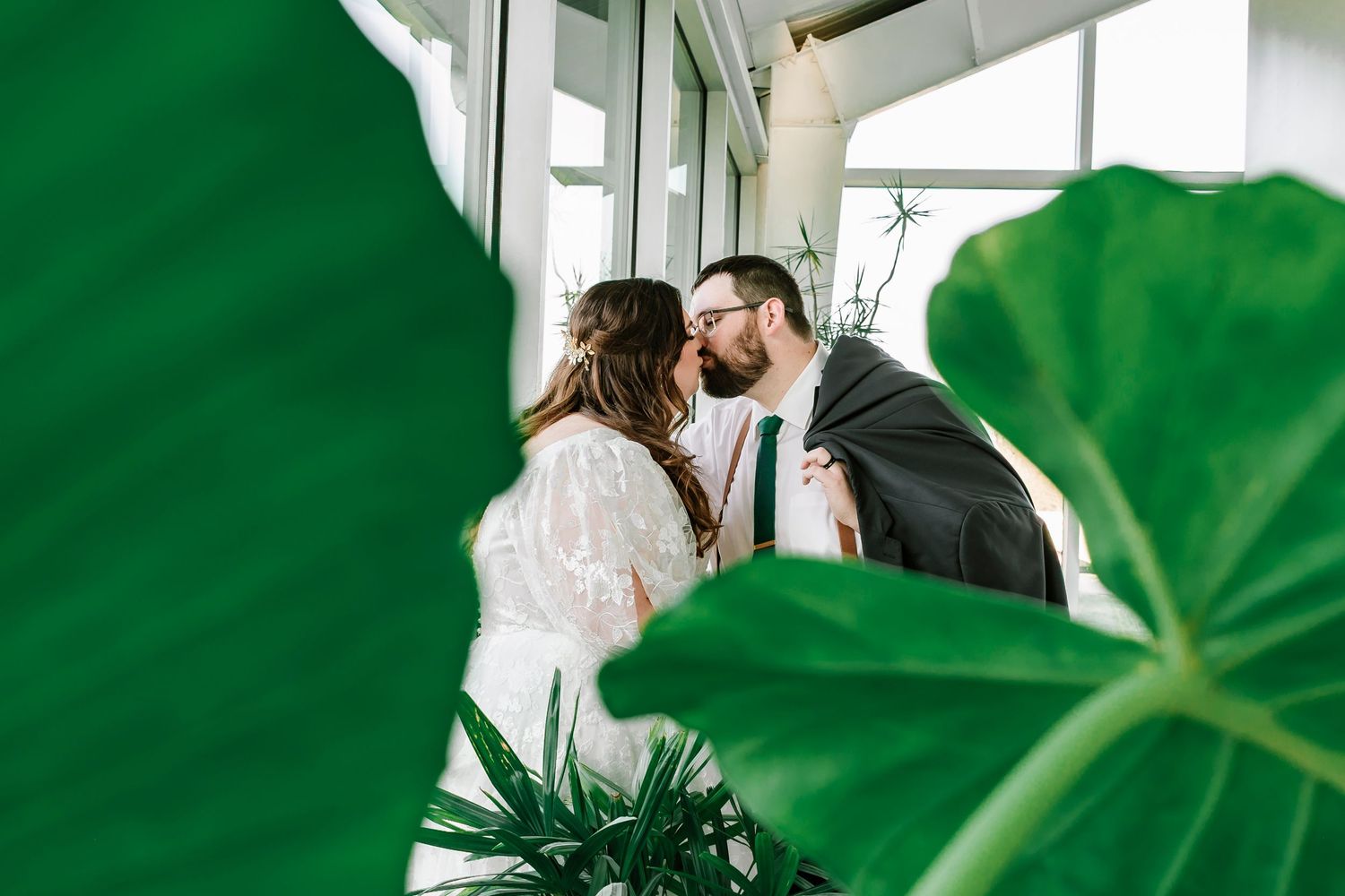 Two people share an intimate moment among lush green foliage in a bright indoor setting.