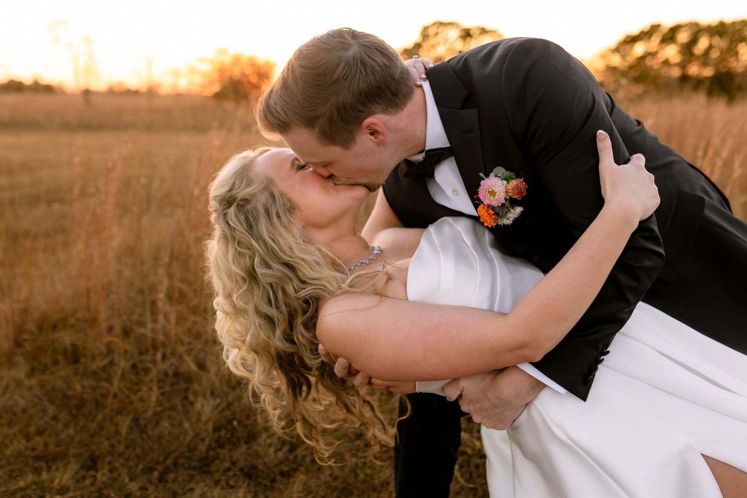 Bride and groom share romantic kiss during golden hour sunset in rustic field with dramatic lighting.