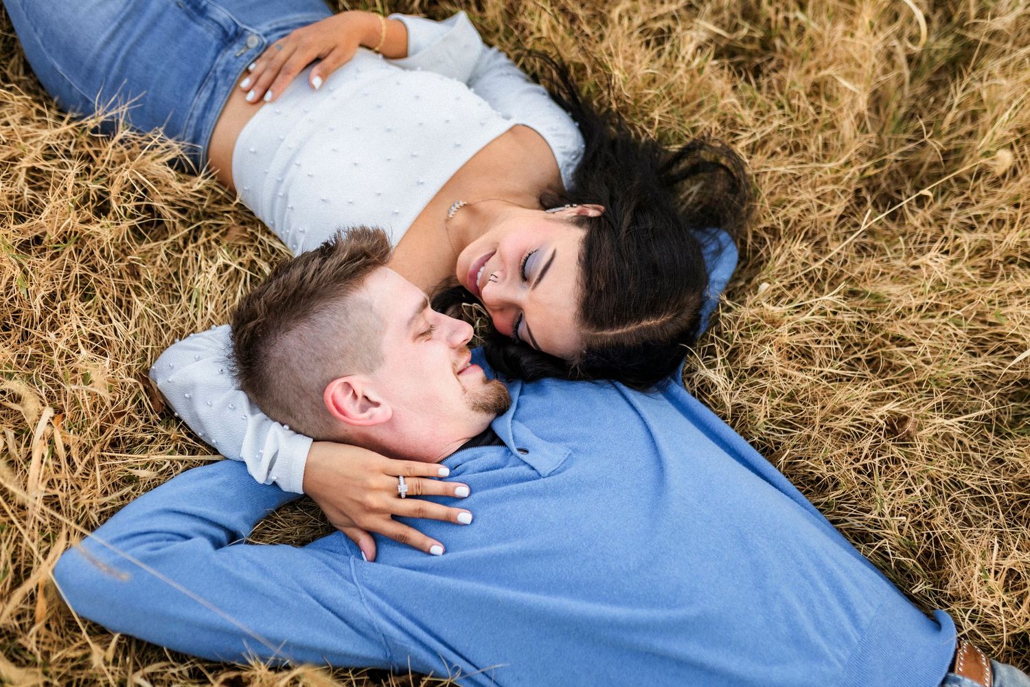 Couple in blue clothing lying together on dry grass field, sharing a sweet aerial view moment.