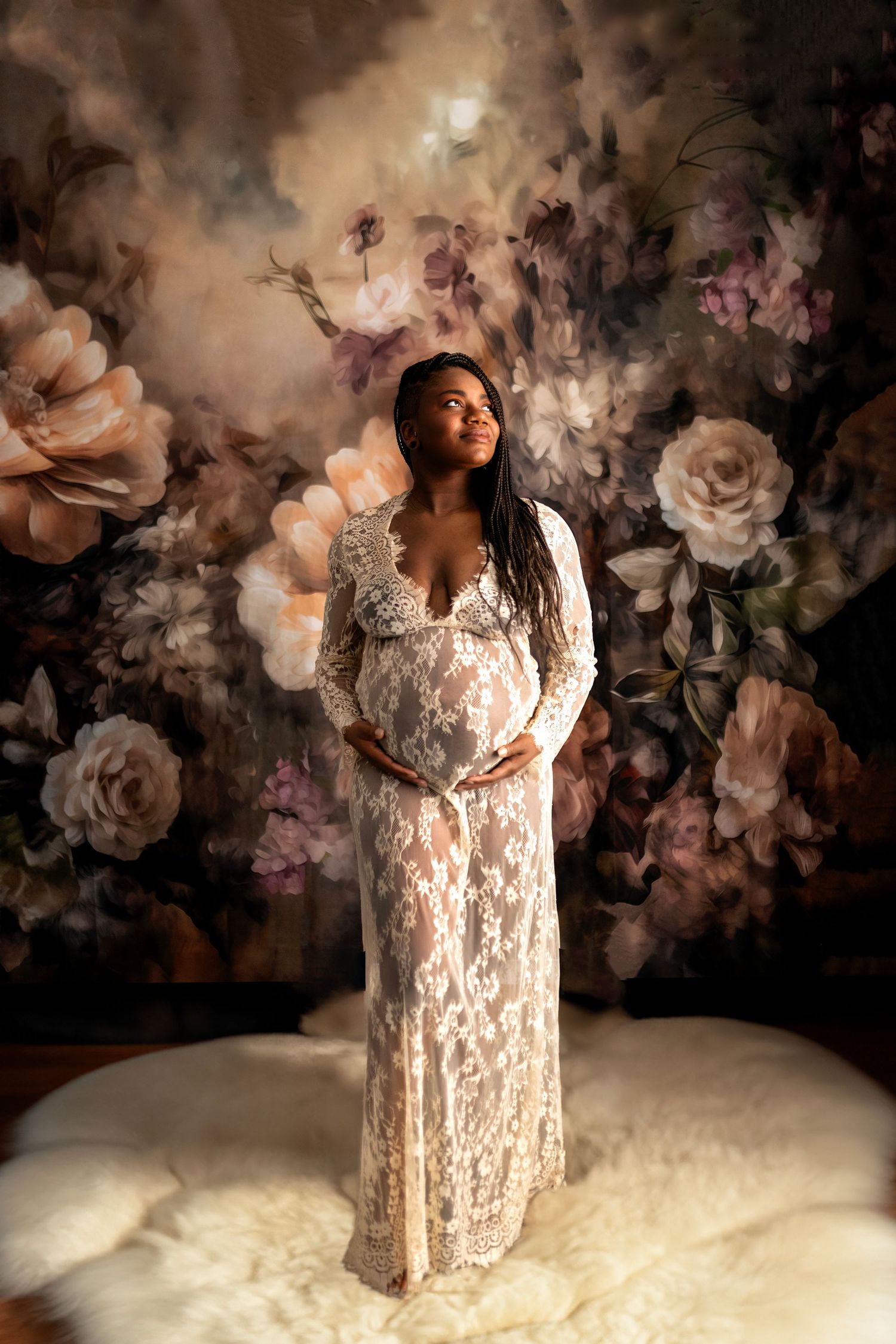 Ethereal maternity photoshoot with white lace gown against dark floral backdrop in moody lighting.