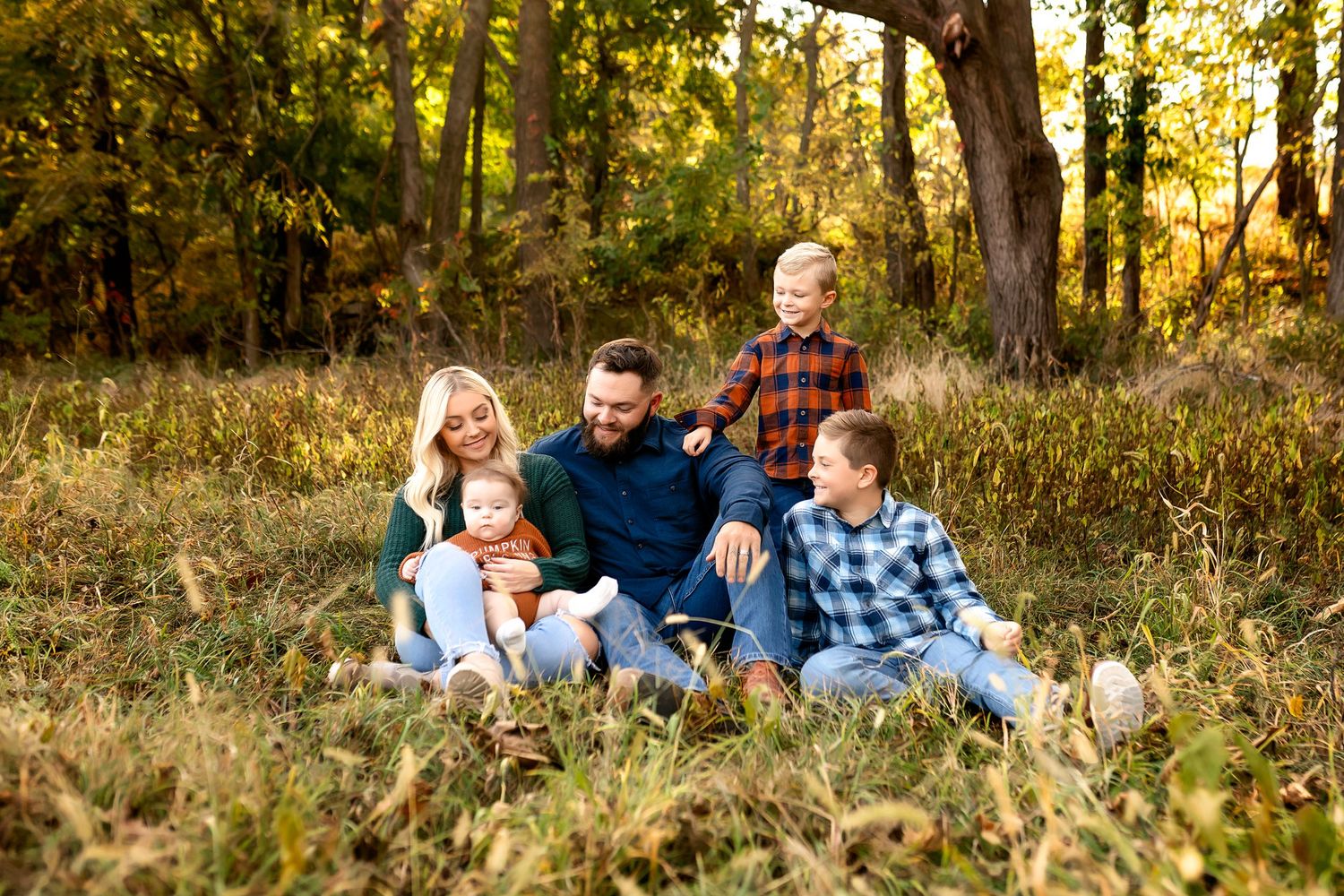 A loving family relaxes together outdoors in a grassy field surrounded by autumn trees at golden hour.
