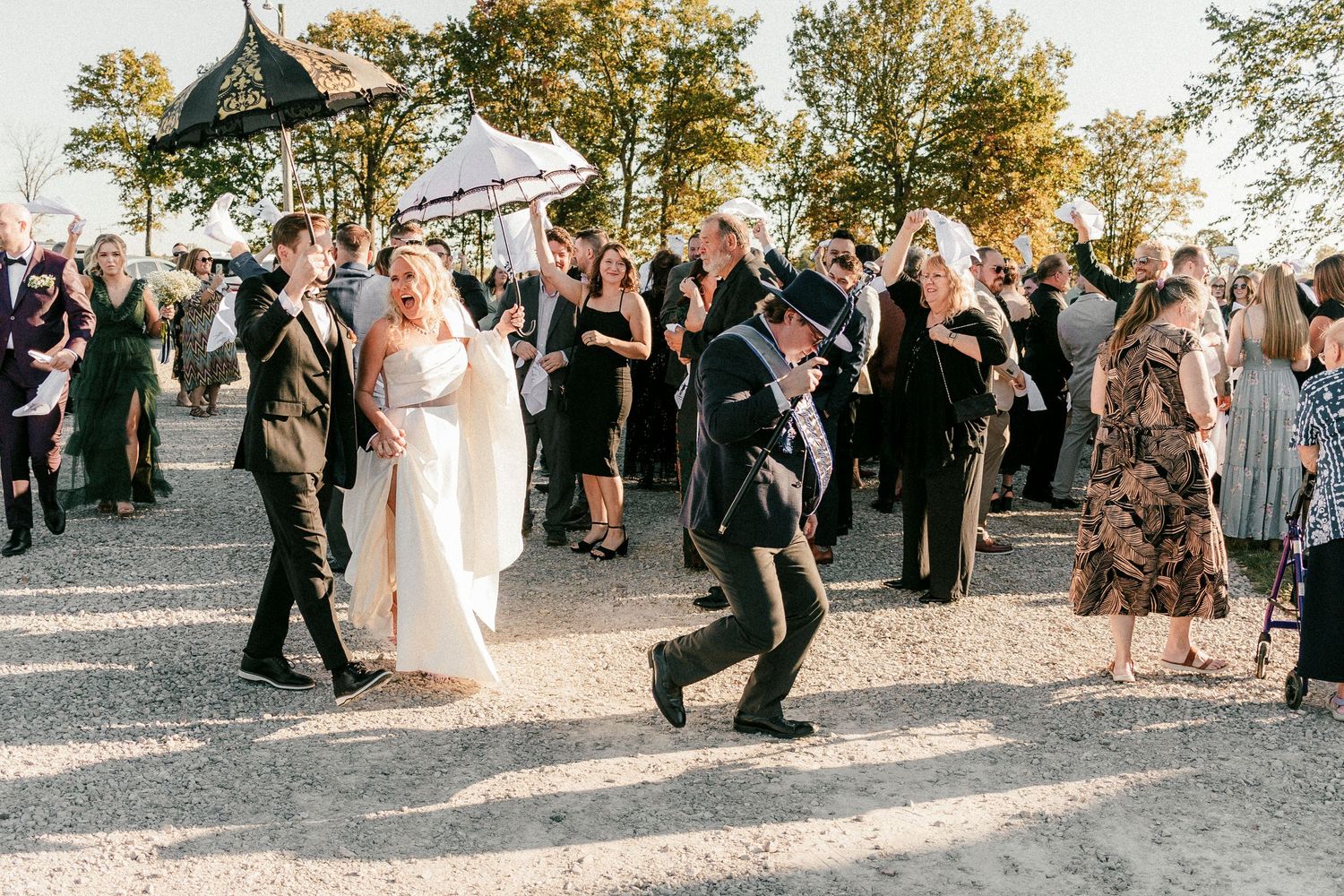 Wedding party celebrates with guests walking down outdoor path during golden hour while child jumps playfully in foreground.