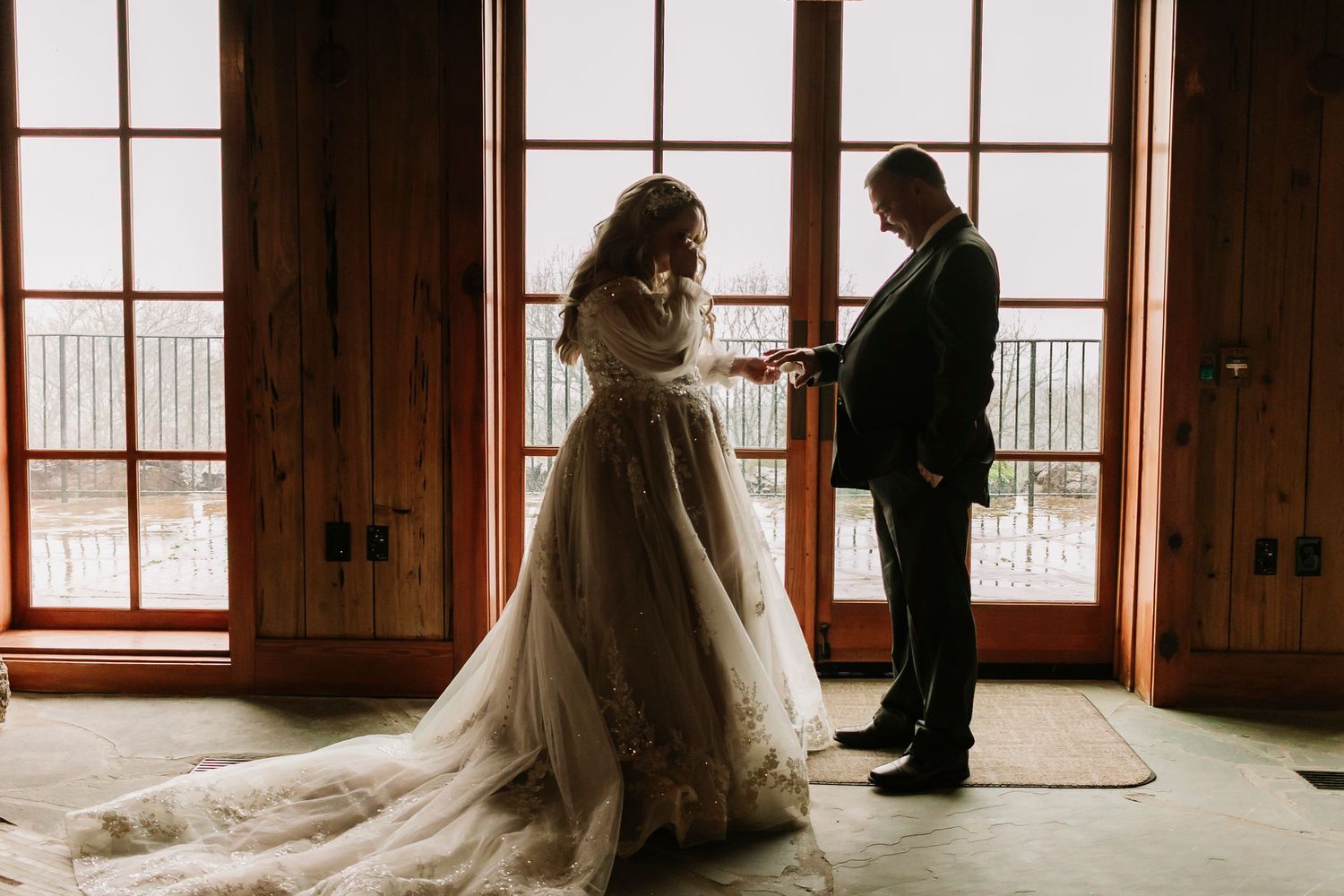 Father and daughter stands by large windows in rustic Top of the Rock Chapel, with the bride wearing a flowing wedding gown with long train.