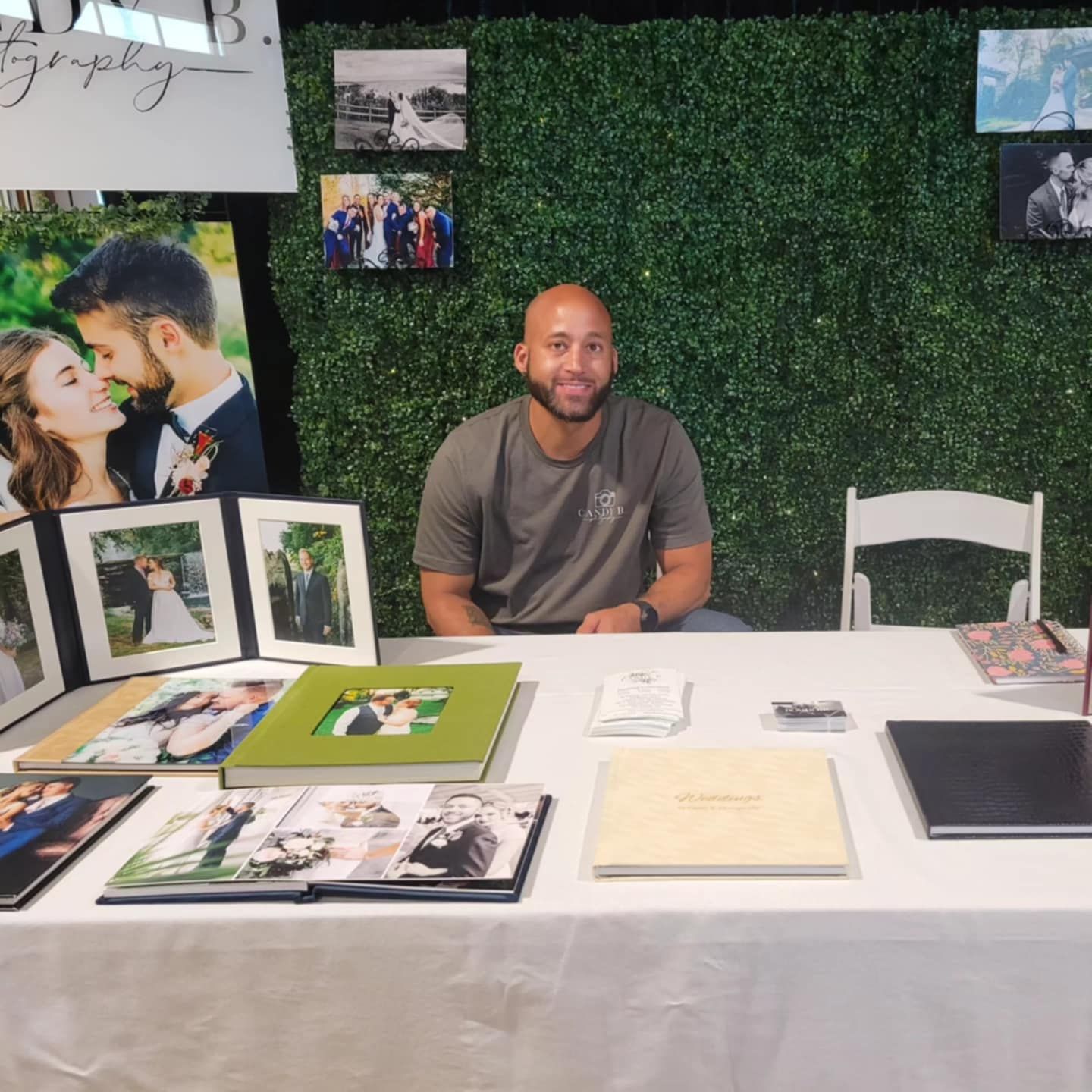 A photographer sits at a display table with photo albums and prints laid out on a white surface against a green hedge wall.
