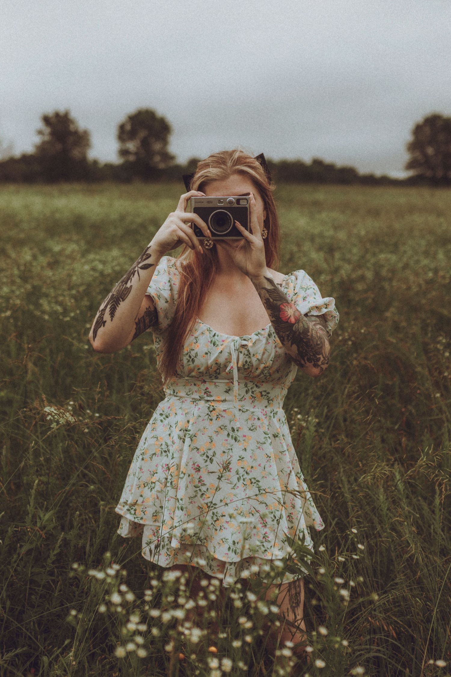 A photographer in a floral dress holds a vintage camera in a wildflower meadow on an overcast day.