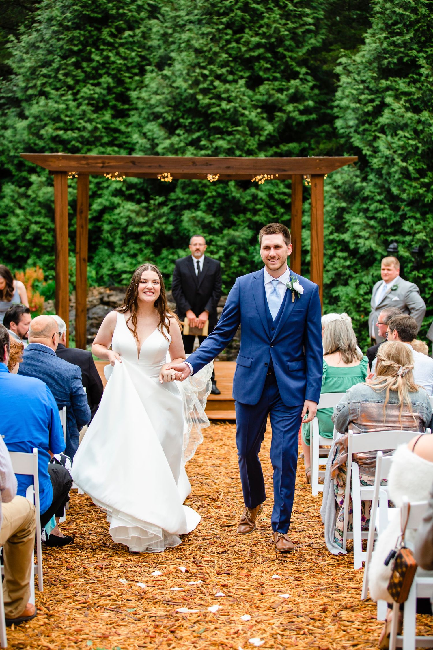 Wedding ceremony setup outdoors with wooden pergola and white chairs on mulch ground surrounded by greenery.