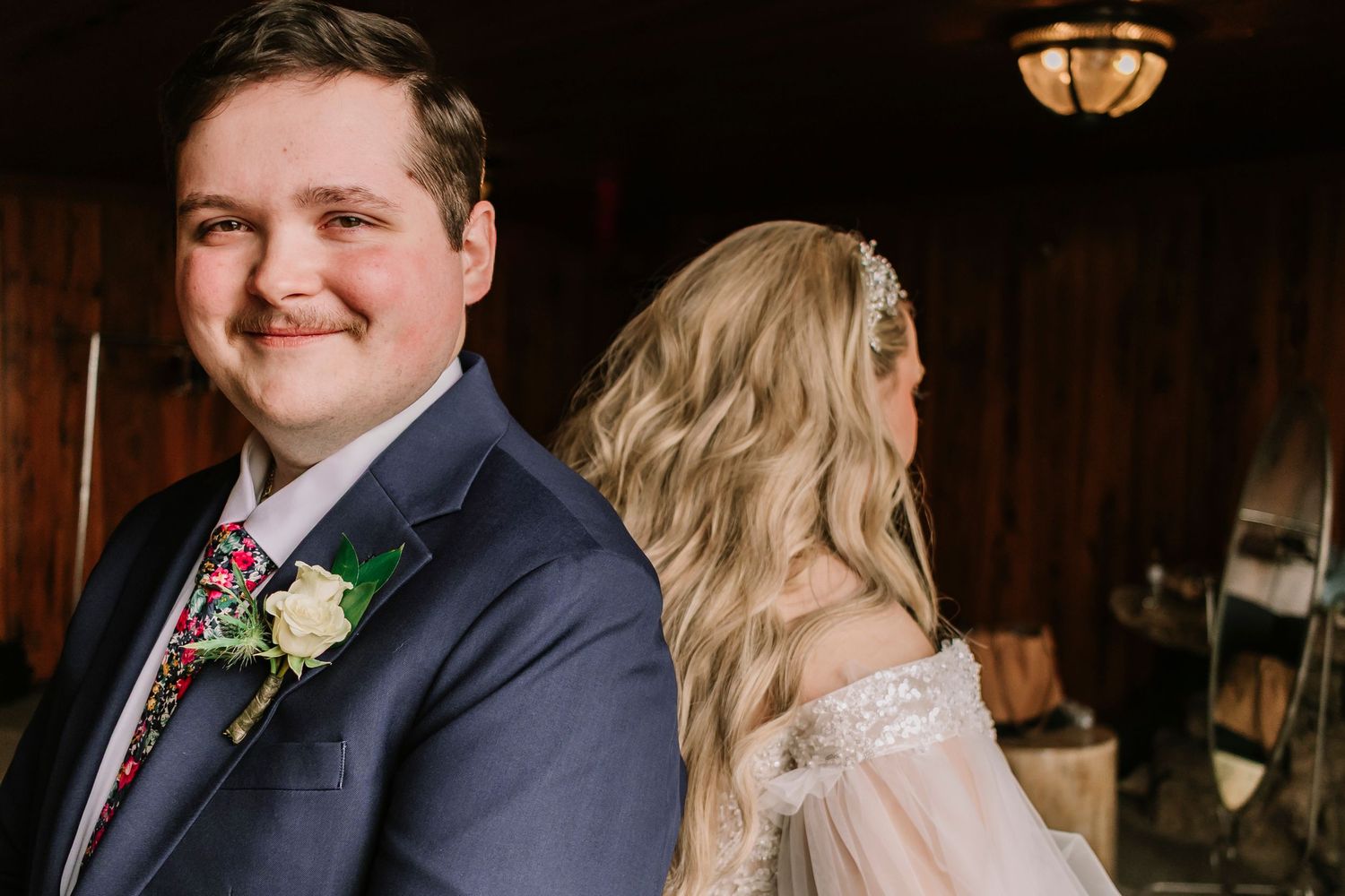 A wedding couple shares a candid first look moment in a rustic wooden venue with warm lighting and boutonniere details.