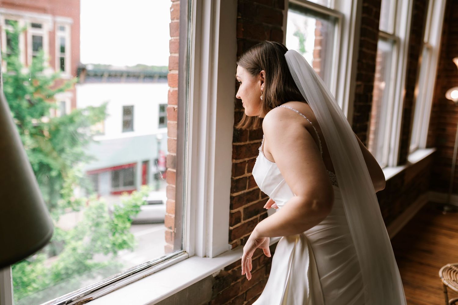 Bride in a strapless white wedding gown gazes thoughtfully out of a large window in airbnb on commercial street in downtown Springfield MO
