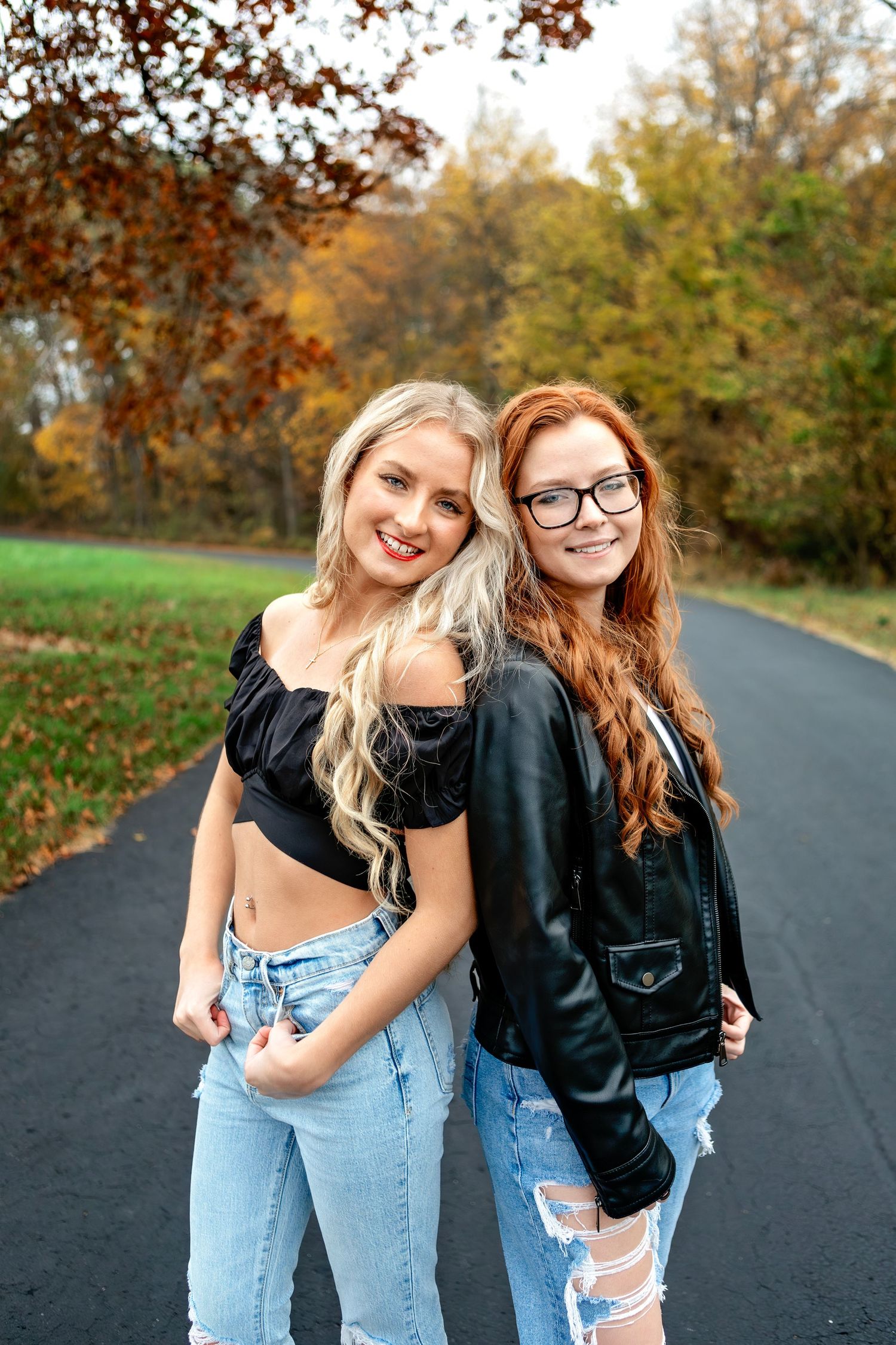 Two sisters in casual outfits pose together on a Springfield Missouri road during autumn with fall foliage in the background.