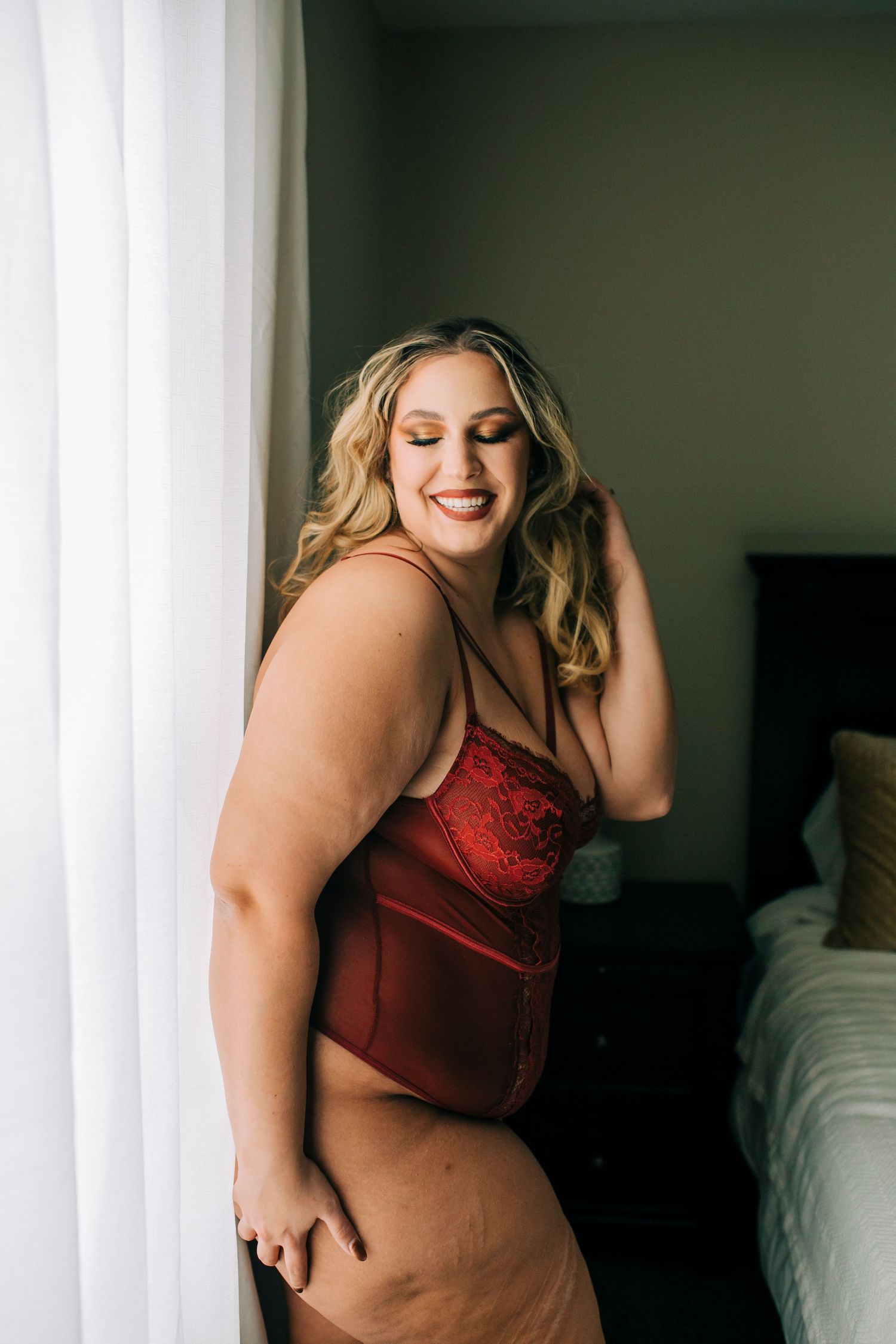 Empowering plus-size boudoir portrait of woman laughing in red bodysuit.