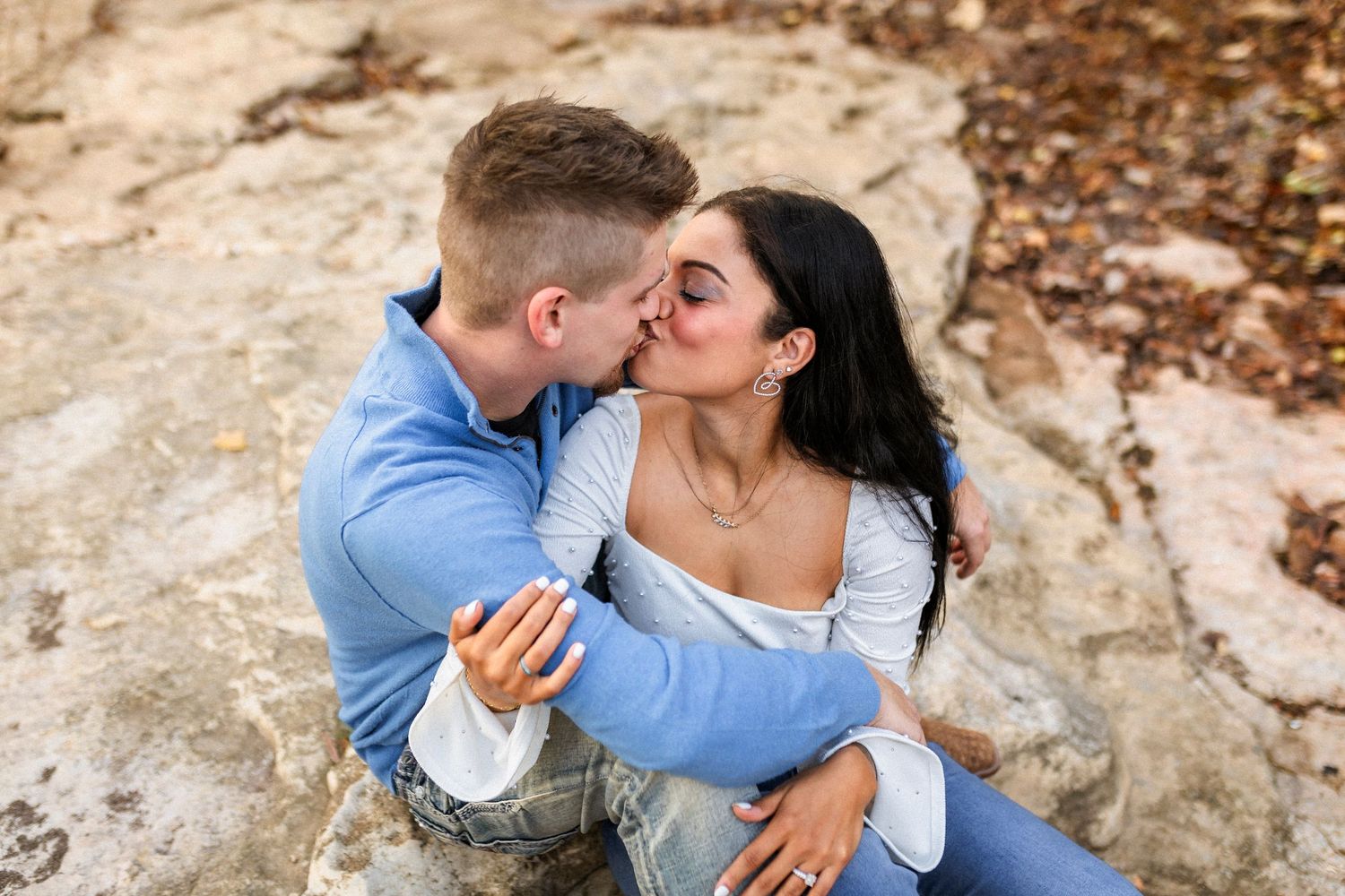 Two people share an intimate romantic moment while sitting together on large rocks outdoors during an engagement photoshoot.