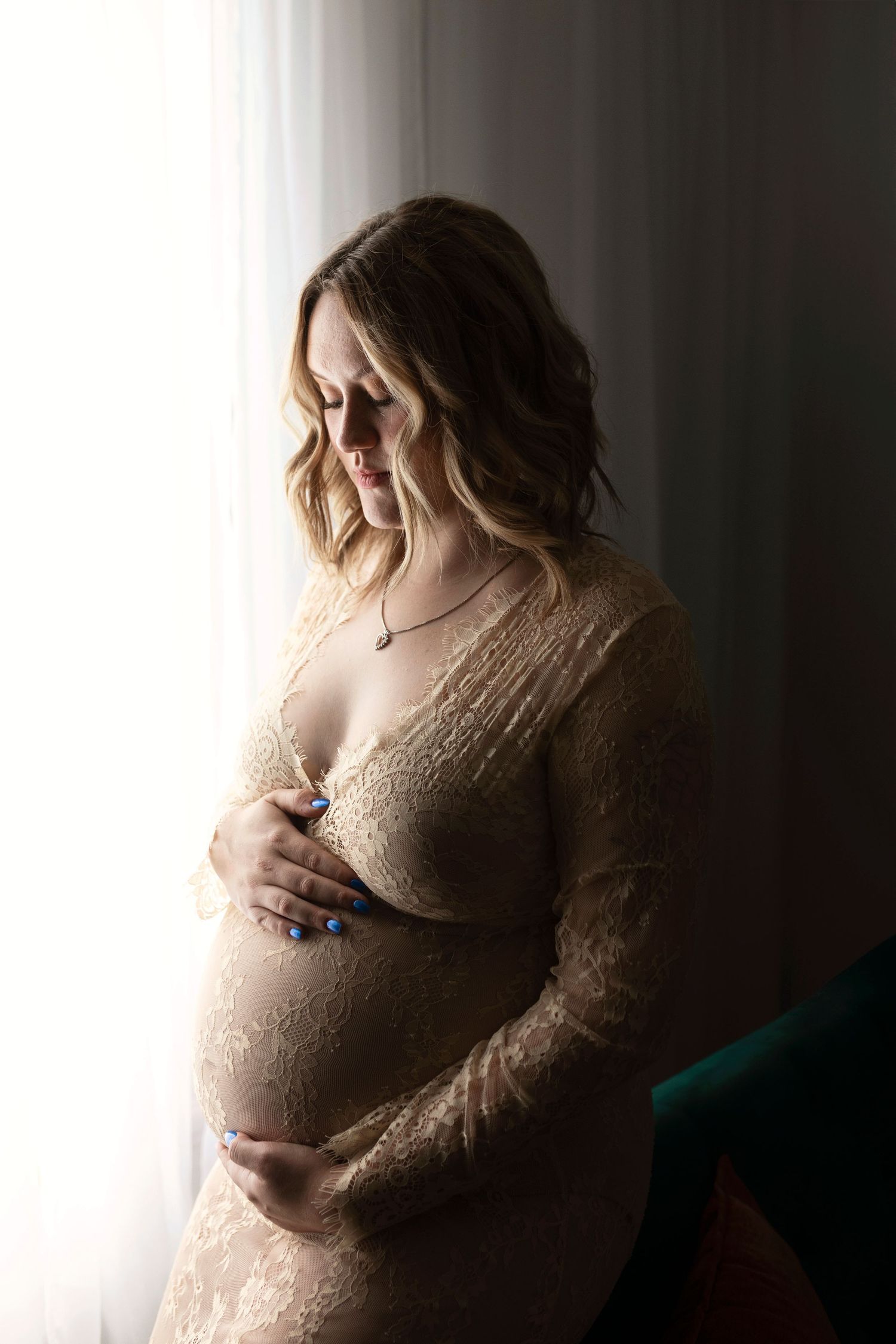A silhouetted pregnant woman in a soft beige sweater stands by a window with natural light casting a gentle glow.