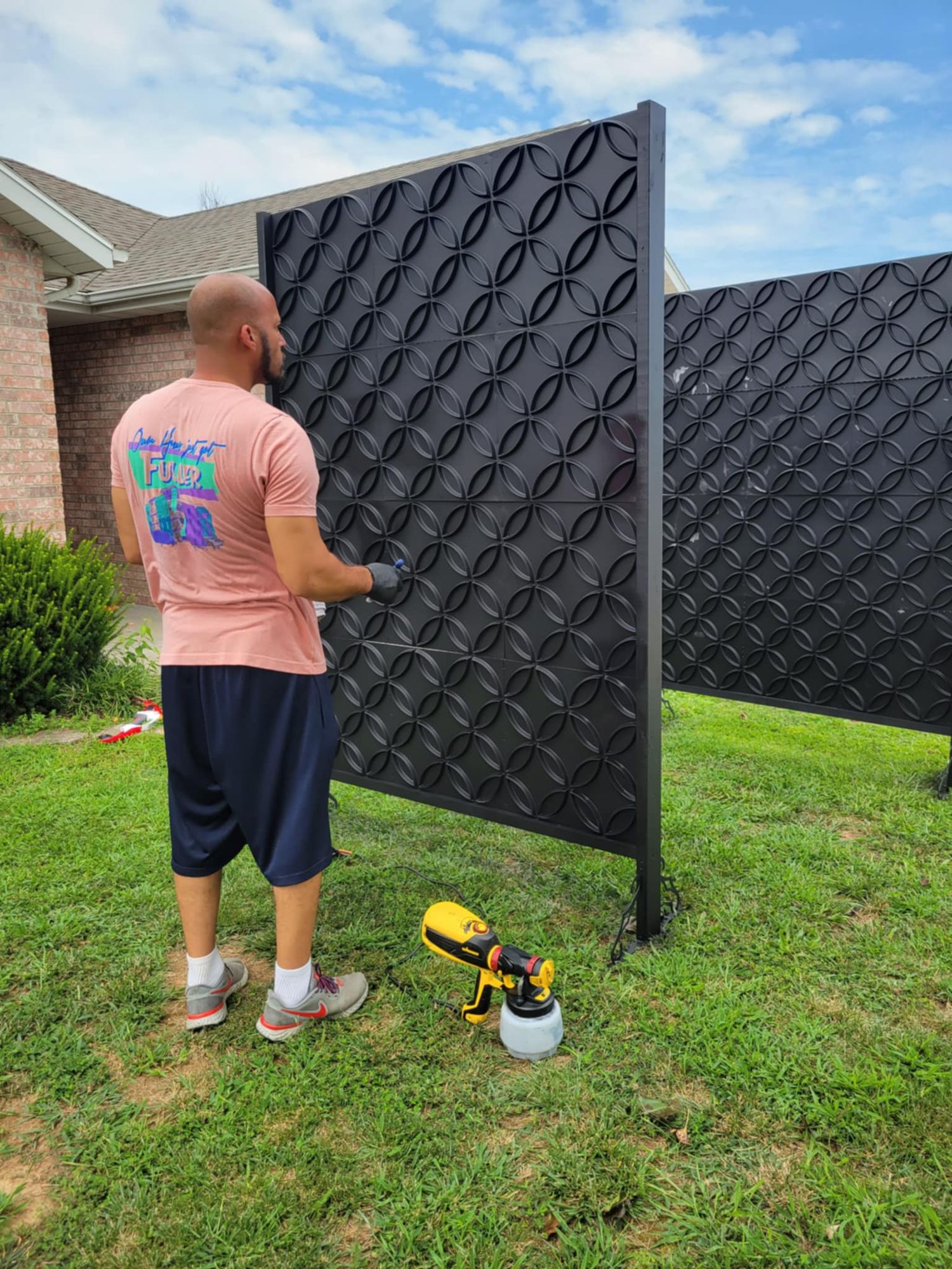Person in pink shirt painting a large dark decorative panel fence in their backyard on a sunny day.