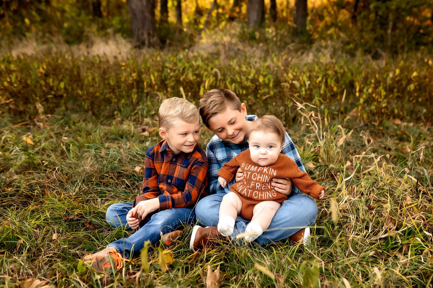 Three children sit close together in tall grass during autumn wearing cozy plaid and knit sweaters.