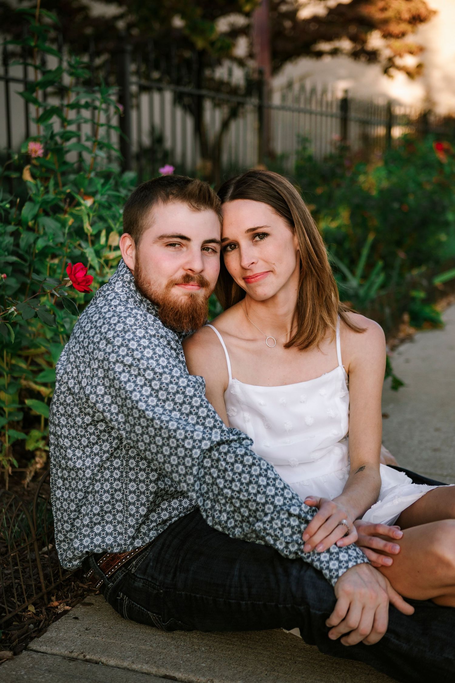 Romantic couple in casual attire lounging together at sunset in a garden setting with wrought iron fence.