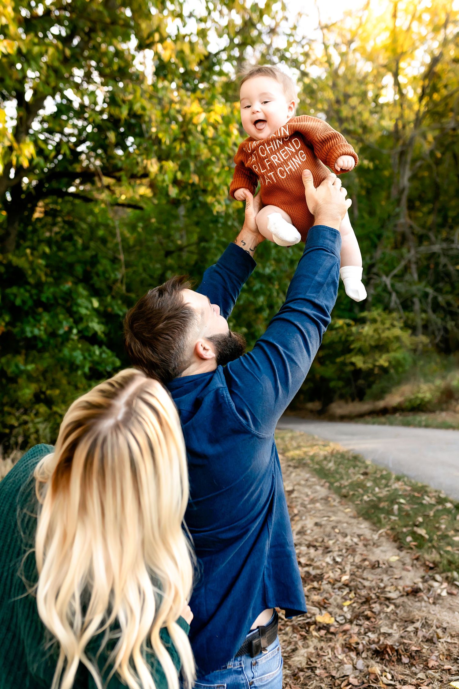 A loving family moment outdoors as joyful smiles are shared while lifting a baby wearing a brown sweater against autumn foliage.