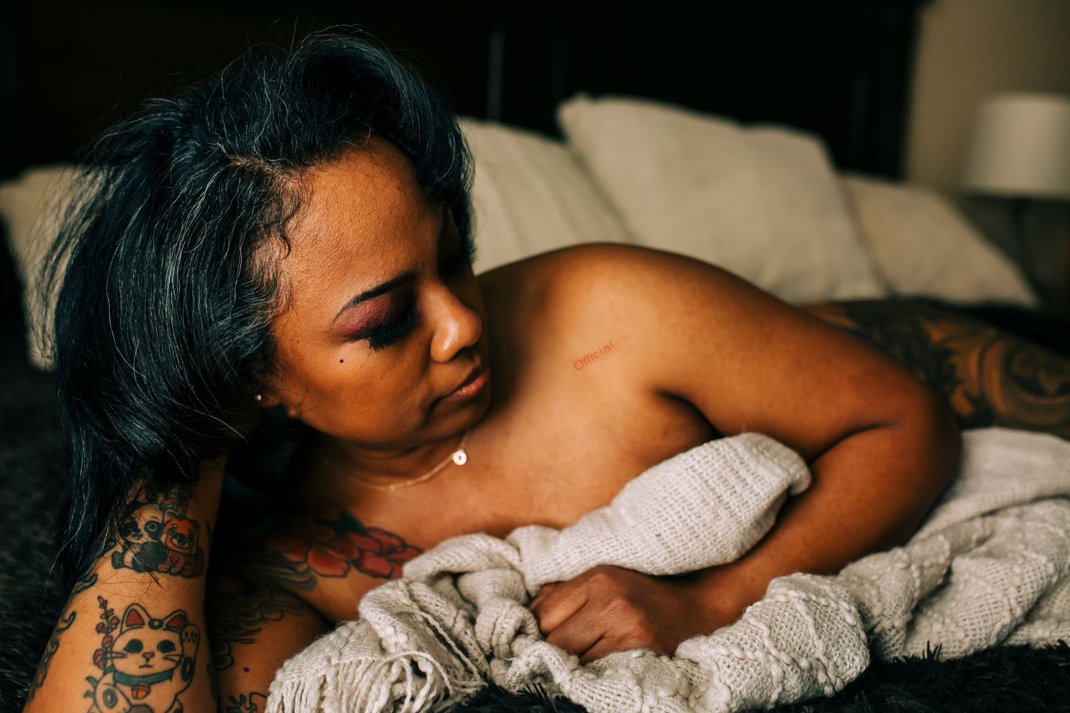 Artistic boudoir photo showing a tattooed woman laying in bed wrapped in white linens.