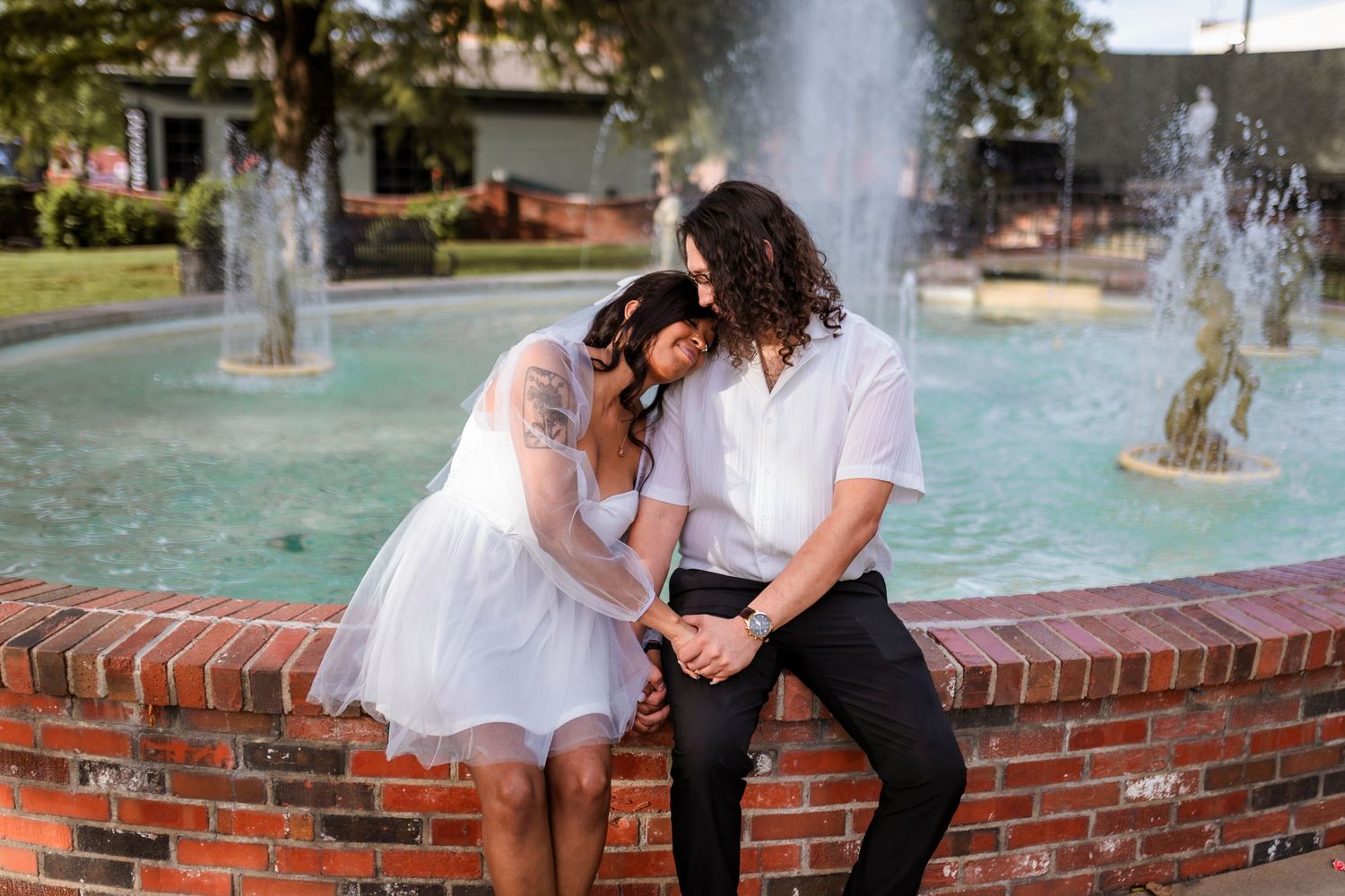 Couple sits intimately on brick fountain ledge with water sprays dancing in background during romantic moment.