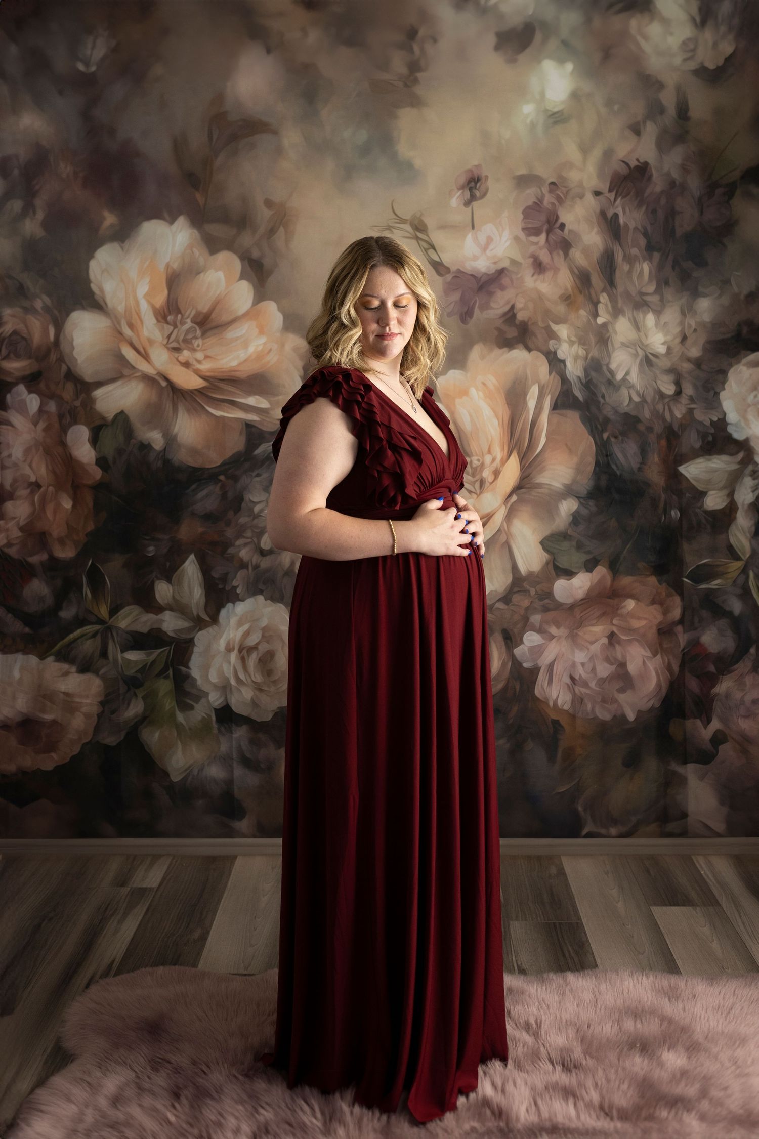 A velvet burgundy maternity gown against a moody floral backdrop with pink roses and dark wood flooring.