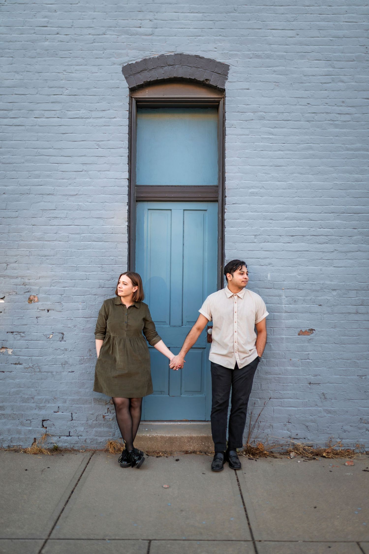 A couple holds hands while standing against a blue painted brick wall with a teal door.
