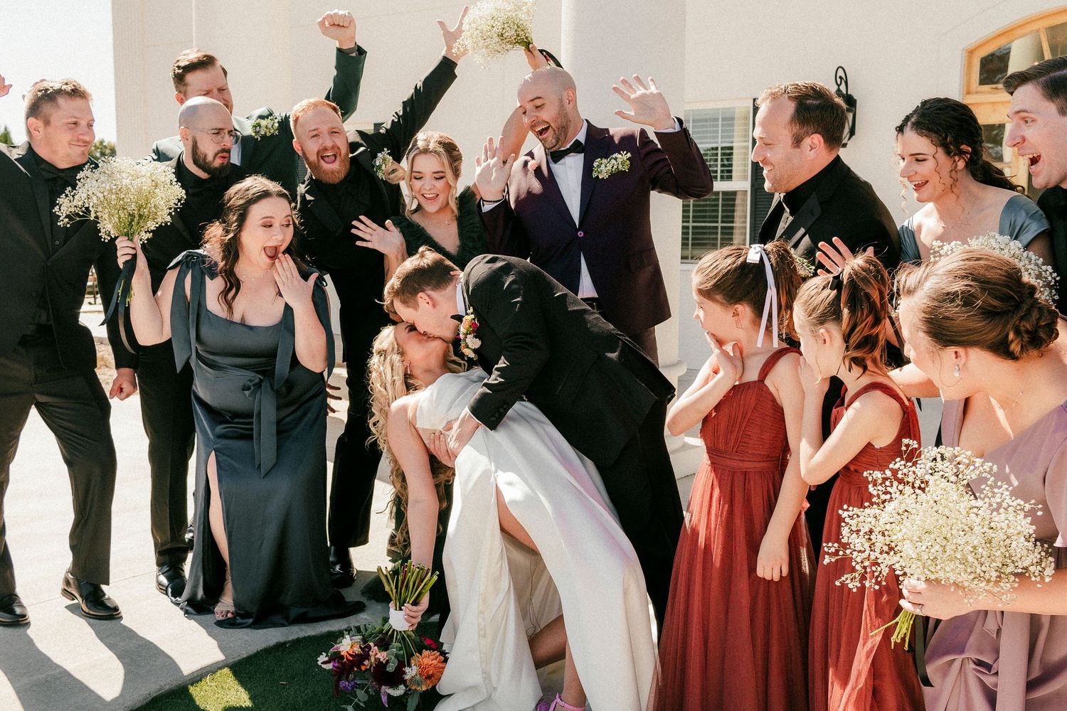 A wedding party celebrates joyfully outdoors, with bridesmaids in burgundy dresses and groomsmen in black suits.