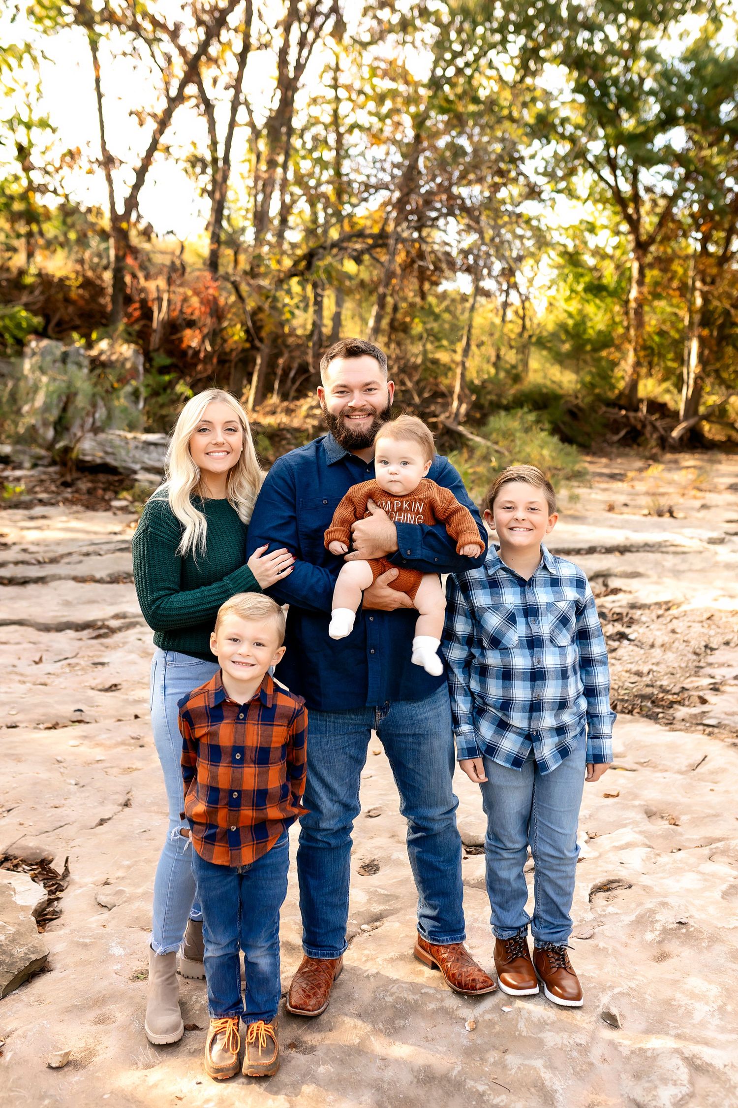 Family wearing coordinated plaid shirts poses together on rocky outdoor terrain at sunset.