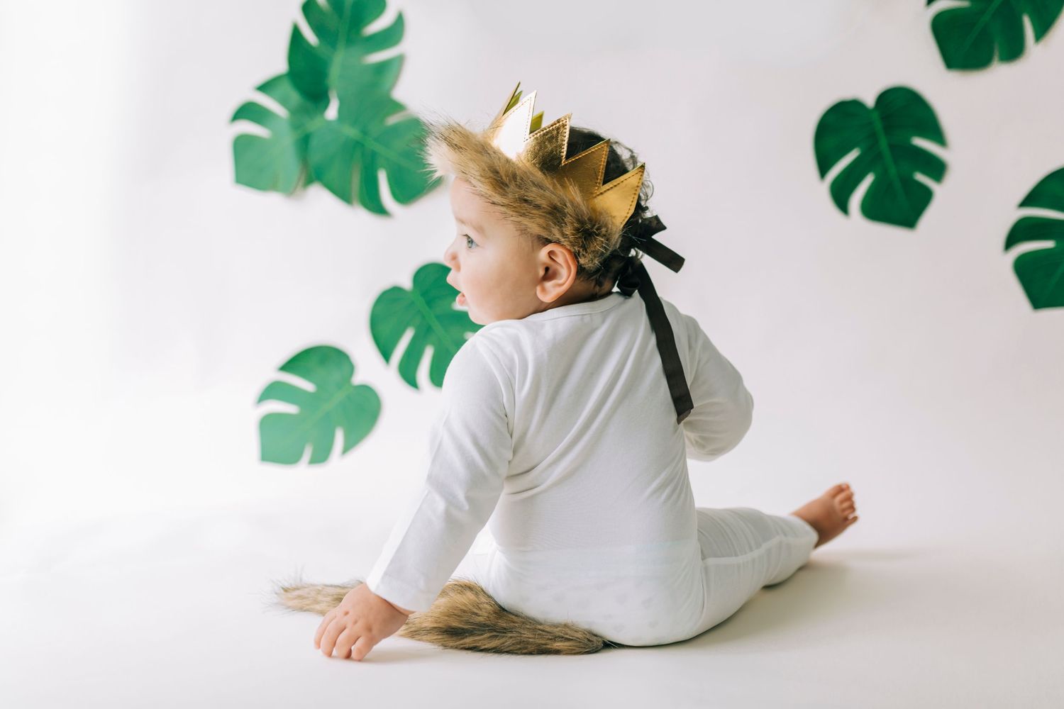 Young child in white outfit sits on floor against white backdrop with green tropical monstera leaf decorations.