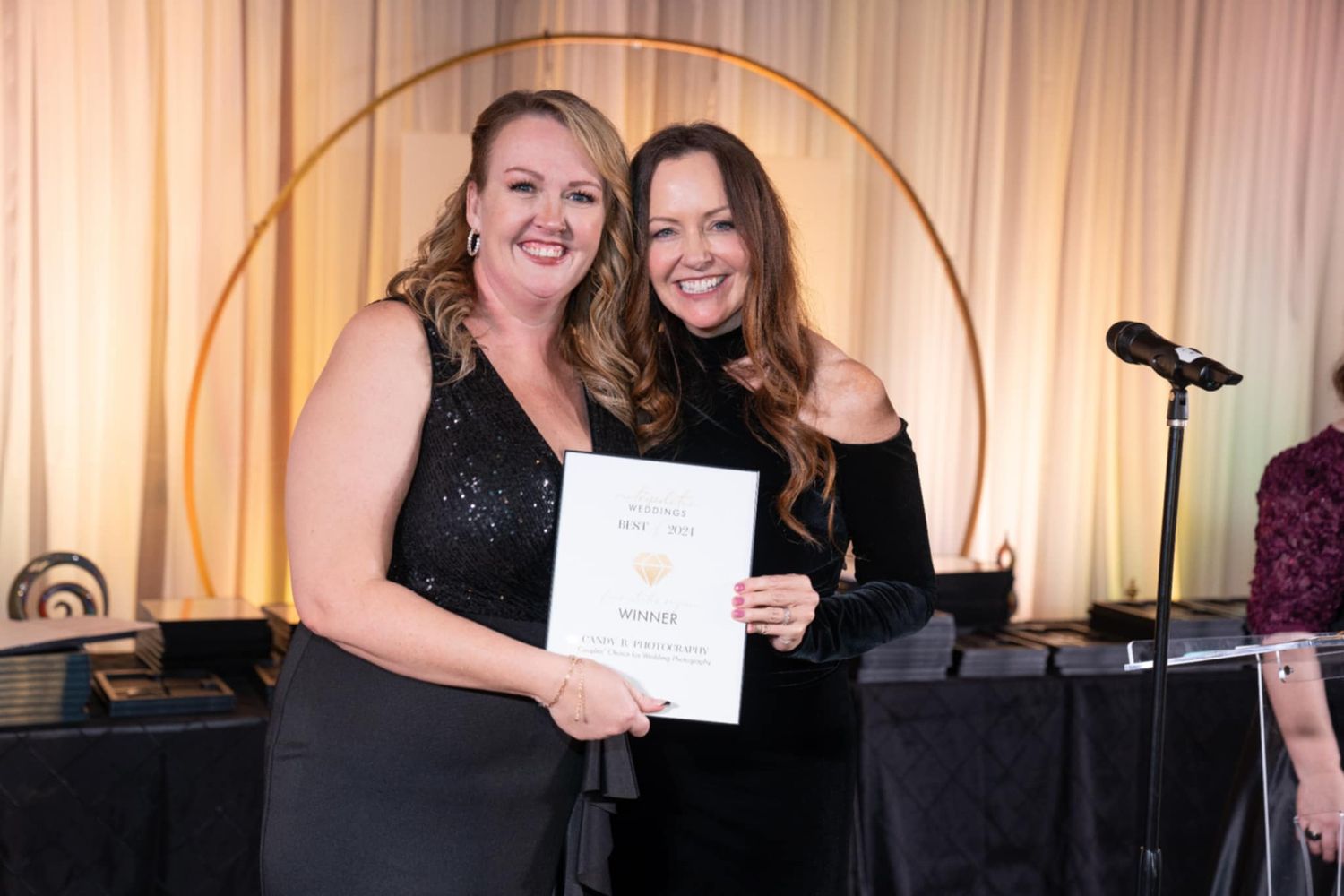 Two people in black dresses pose with a certificate at an award ceremony with stage lighting and microphone visible.