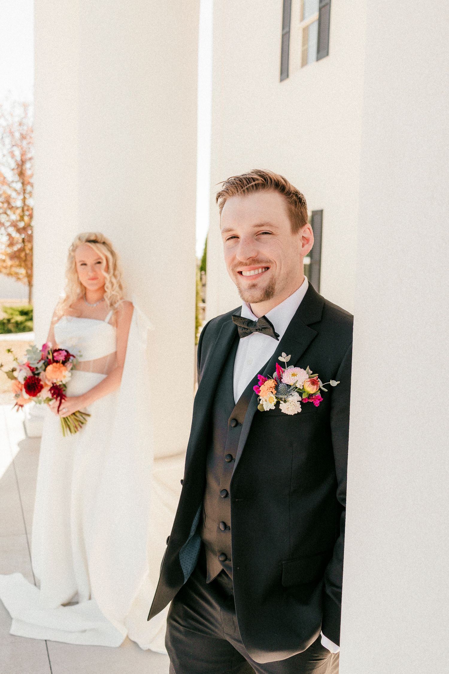 A bride and groom pose separately at a wedding with white columns, fall trees in background, and colorful floral bouquets.
